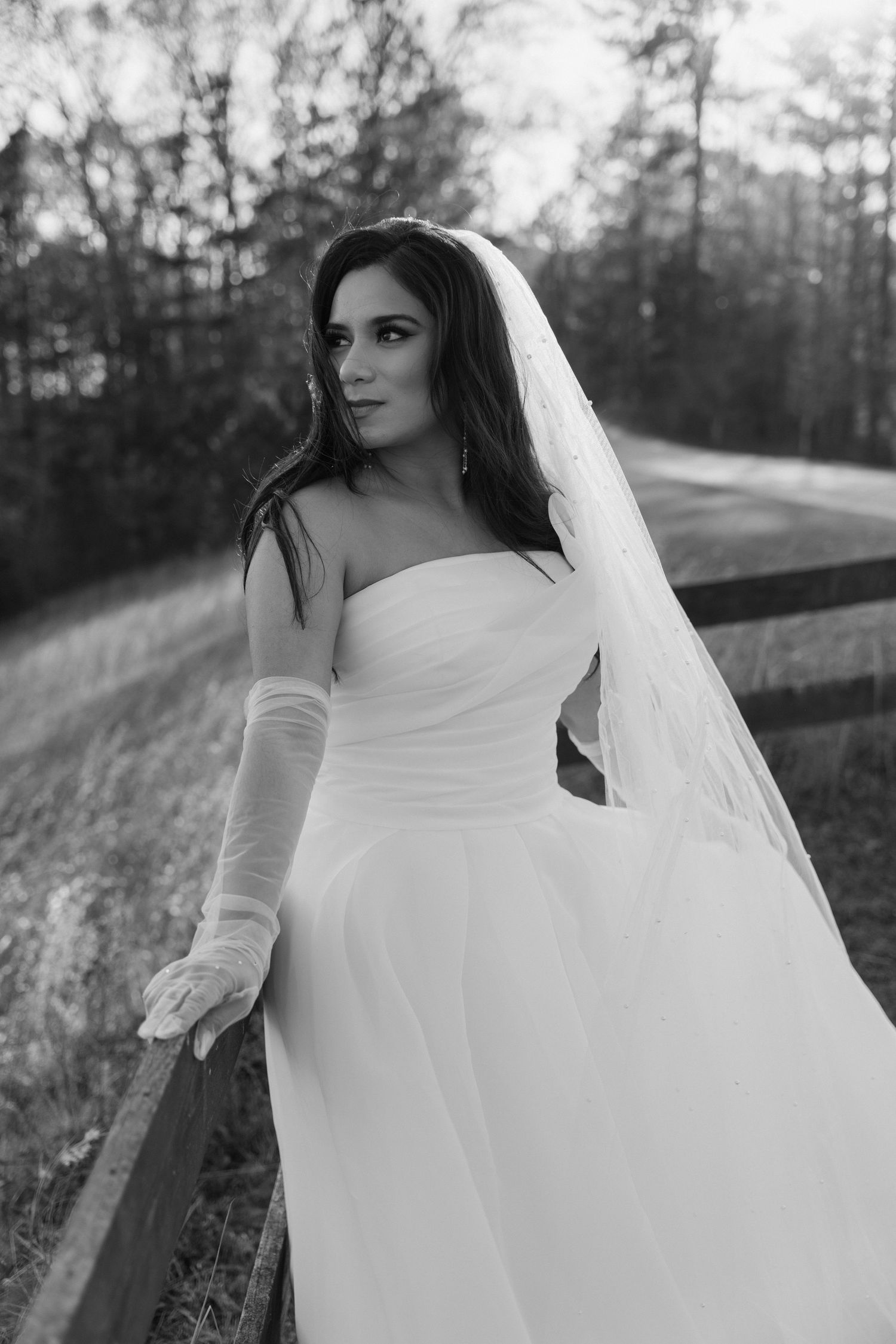 Black and white photo of a bride in a strapless gown, veil, and gloves posing by a fence outdoors.