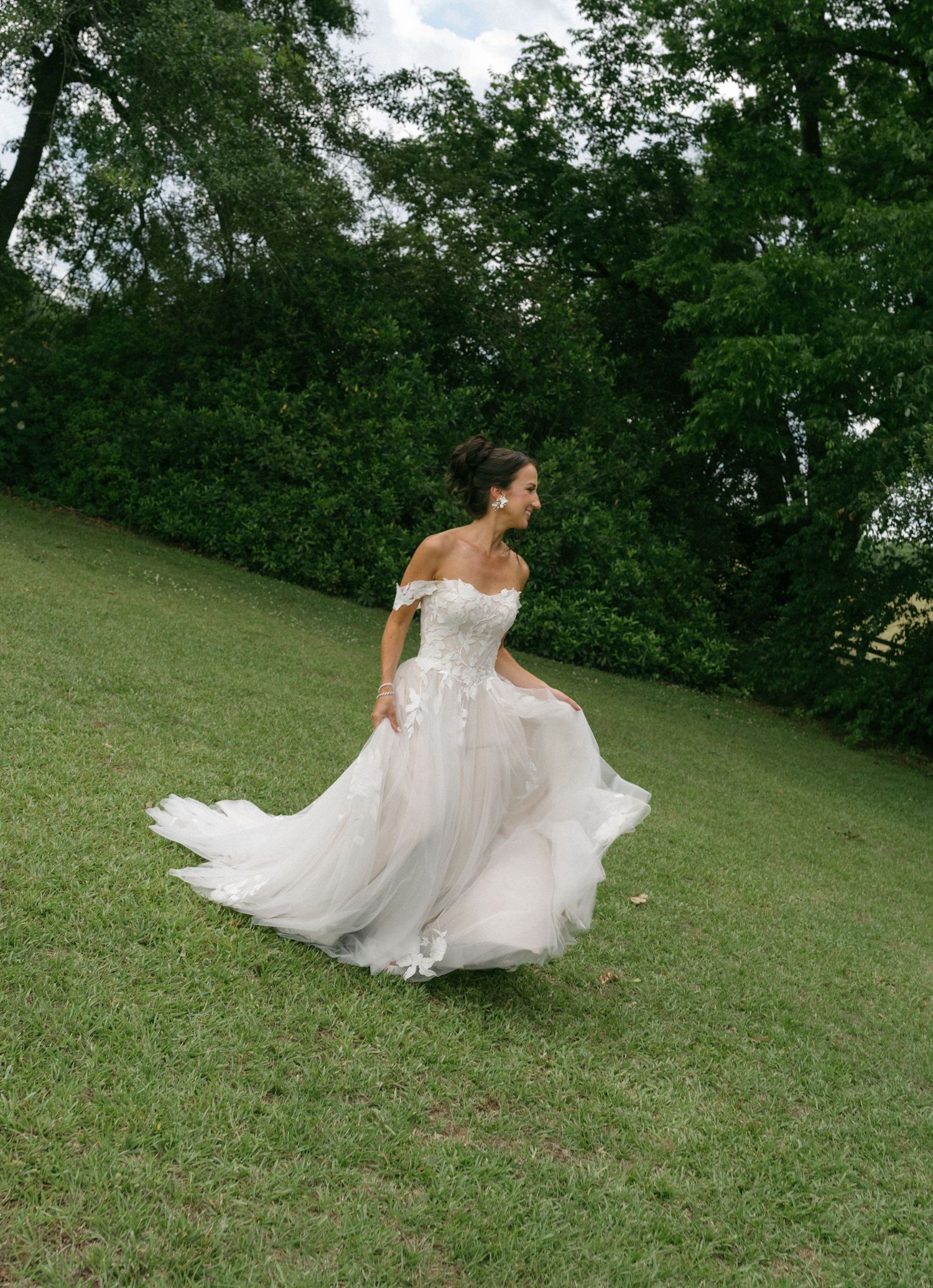 Bride in off-shoulder lace wedding gown twirling on green lawn with lush trees in background.