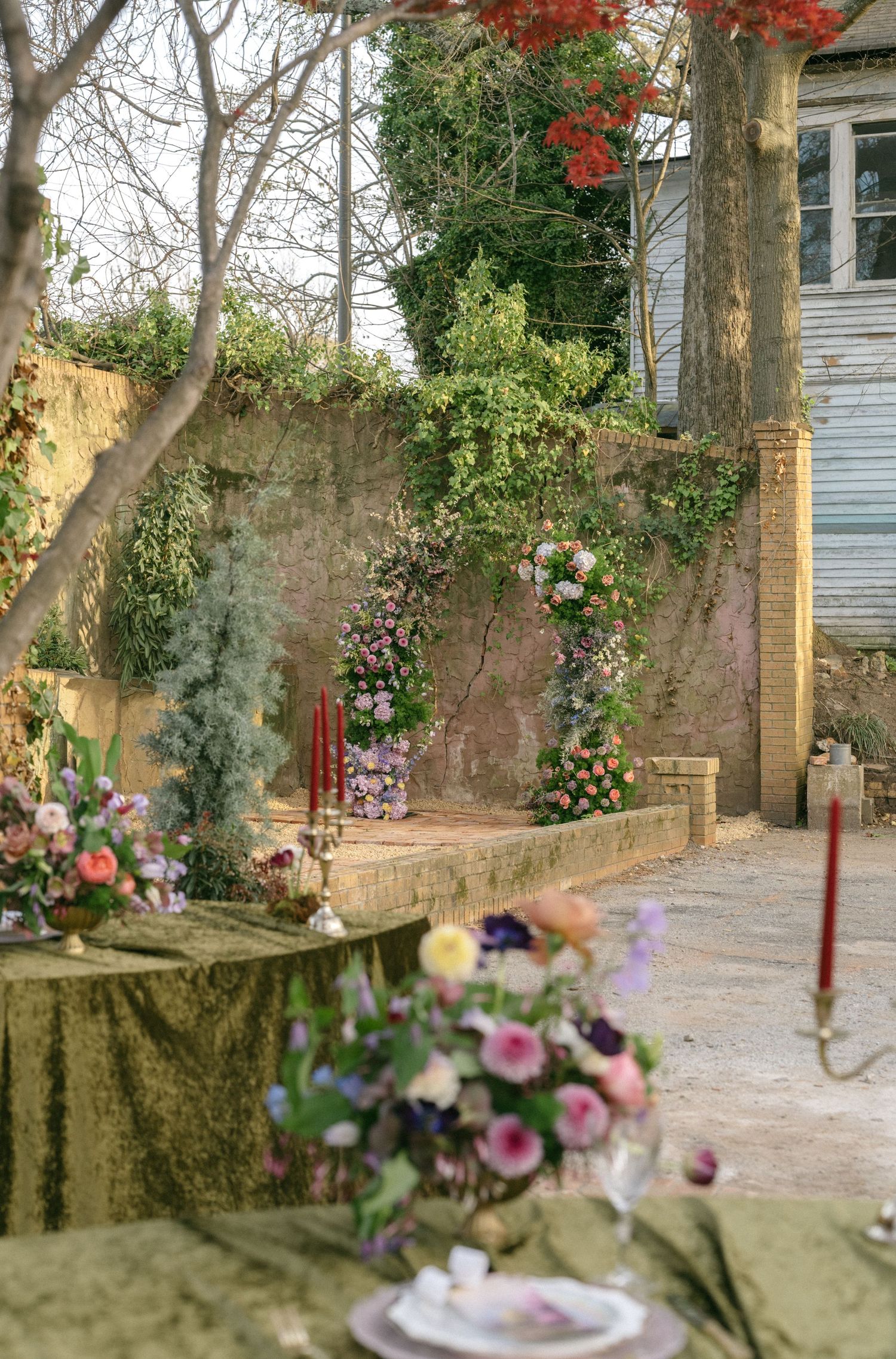 Romantic outdoor wedding reception setup with floral arch, candlesticks, and flower-adorned table in a garden courtyard.