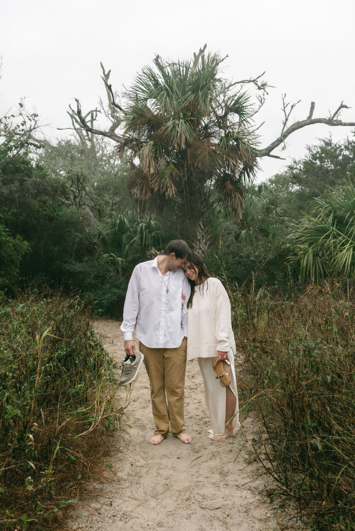 Couple standing barefoot on sandy path surrounded by lush greenery and palm trees, leaning heads together romantically.