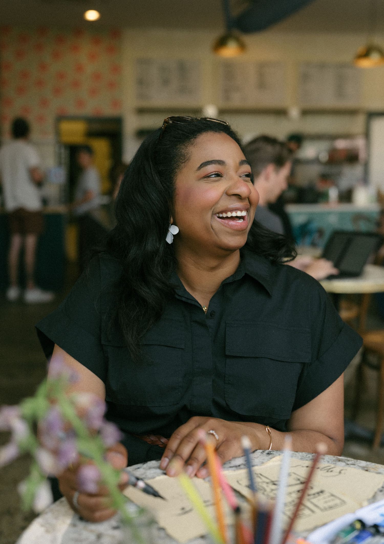 A smiling woman in a black shirt sits at a cafe table with art supplies, laughing joyfully in a bright setting.