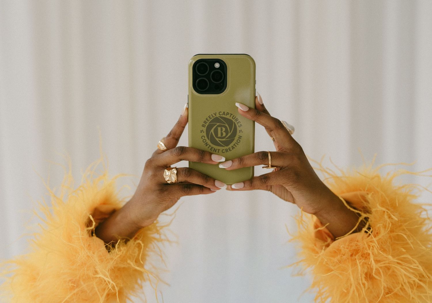 Hands holding a gold phone case with logo, wearing orange feather sleeves and rings.