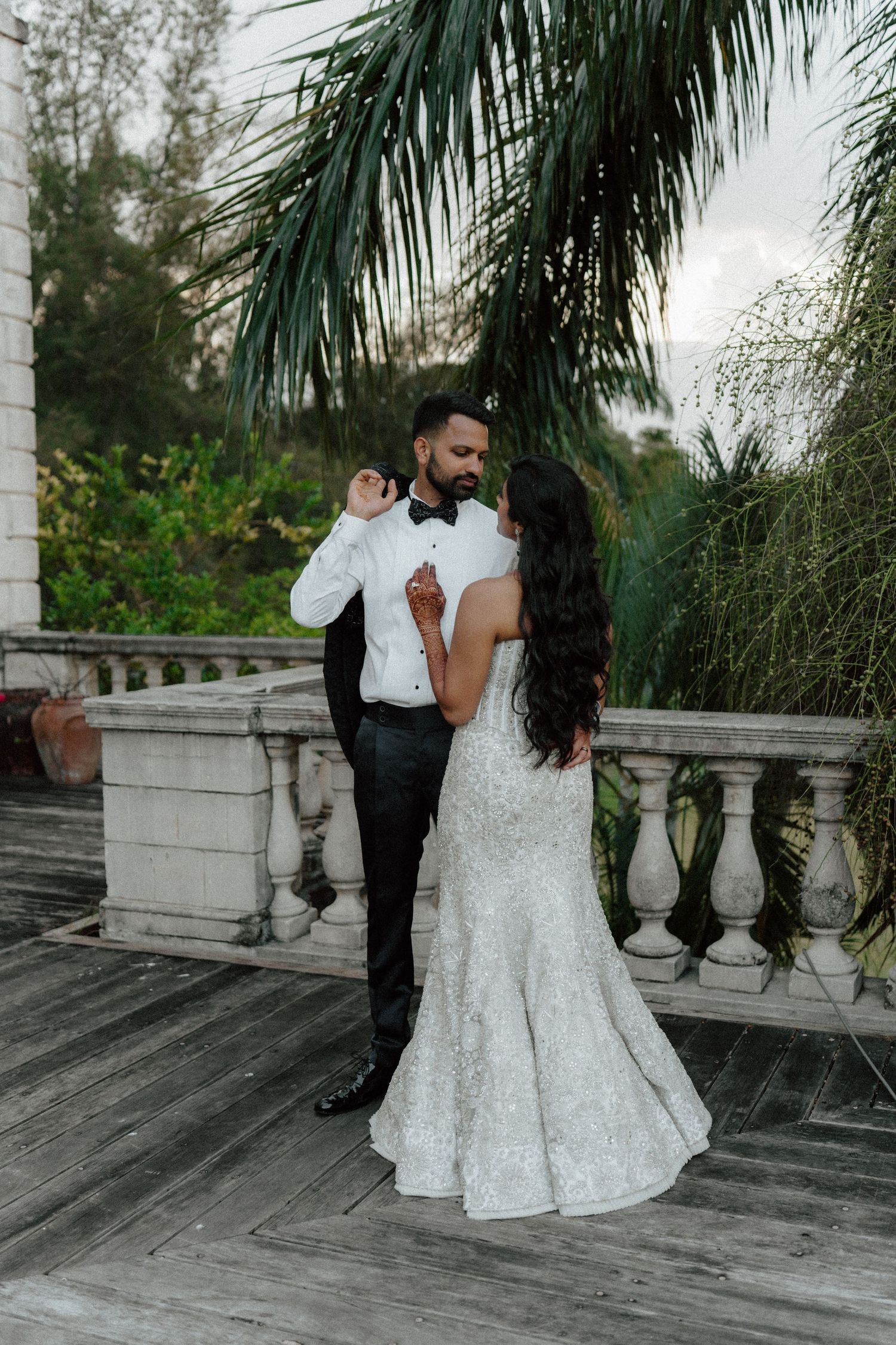 Bride and groom share a romantic moment on an elegant balcony surrounded by lush tropical greenery.