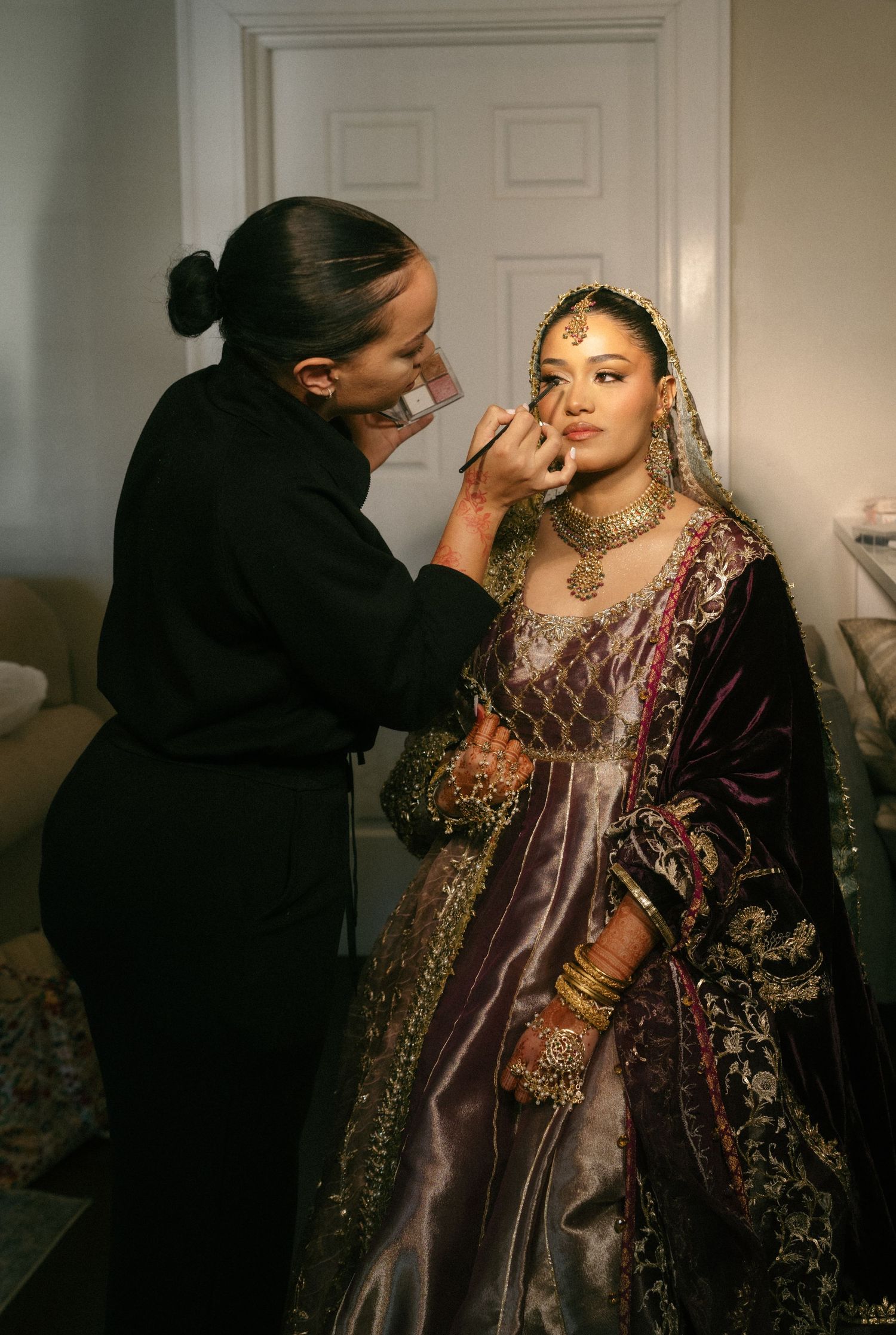 A makeup artist in black applies bridal makeup to a woman in ornate Indian bridal attire with gold jewelry and veil.