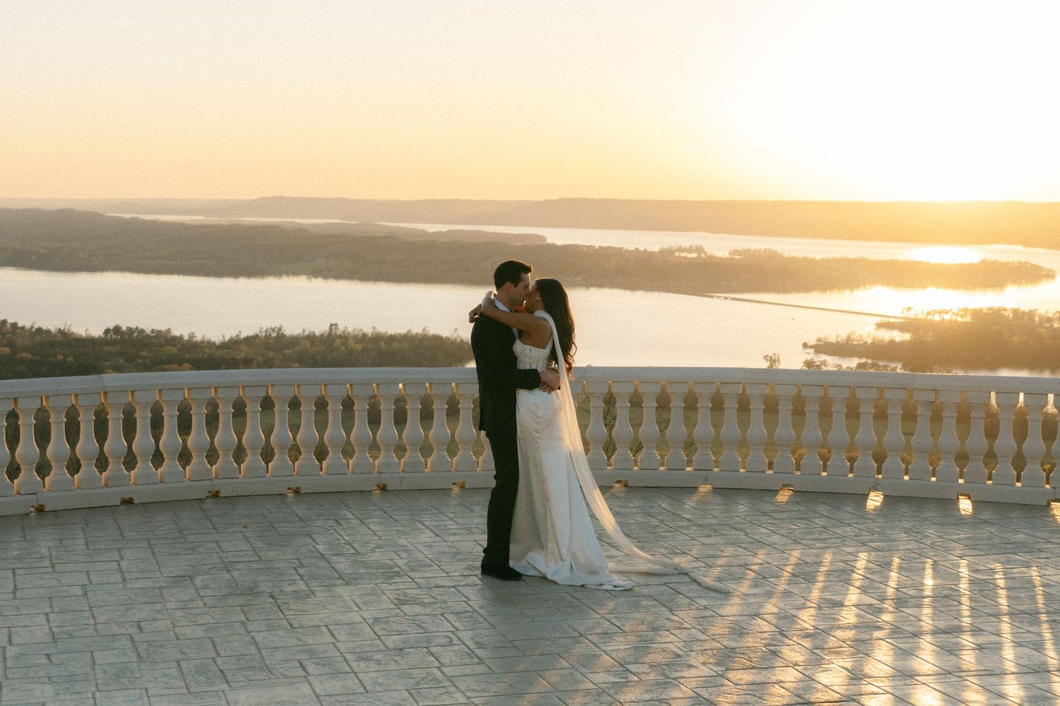 Bride and groom share a romantic kiss on a terrace overlooking a stunning river sunset on their wedding day.