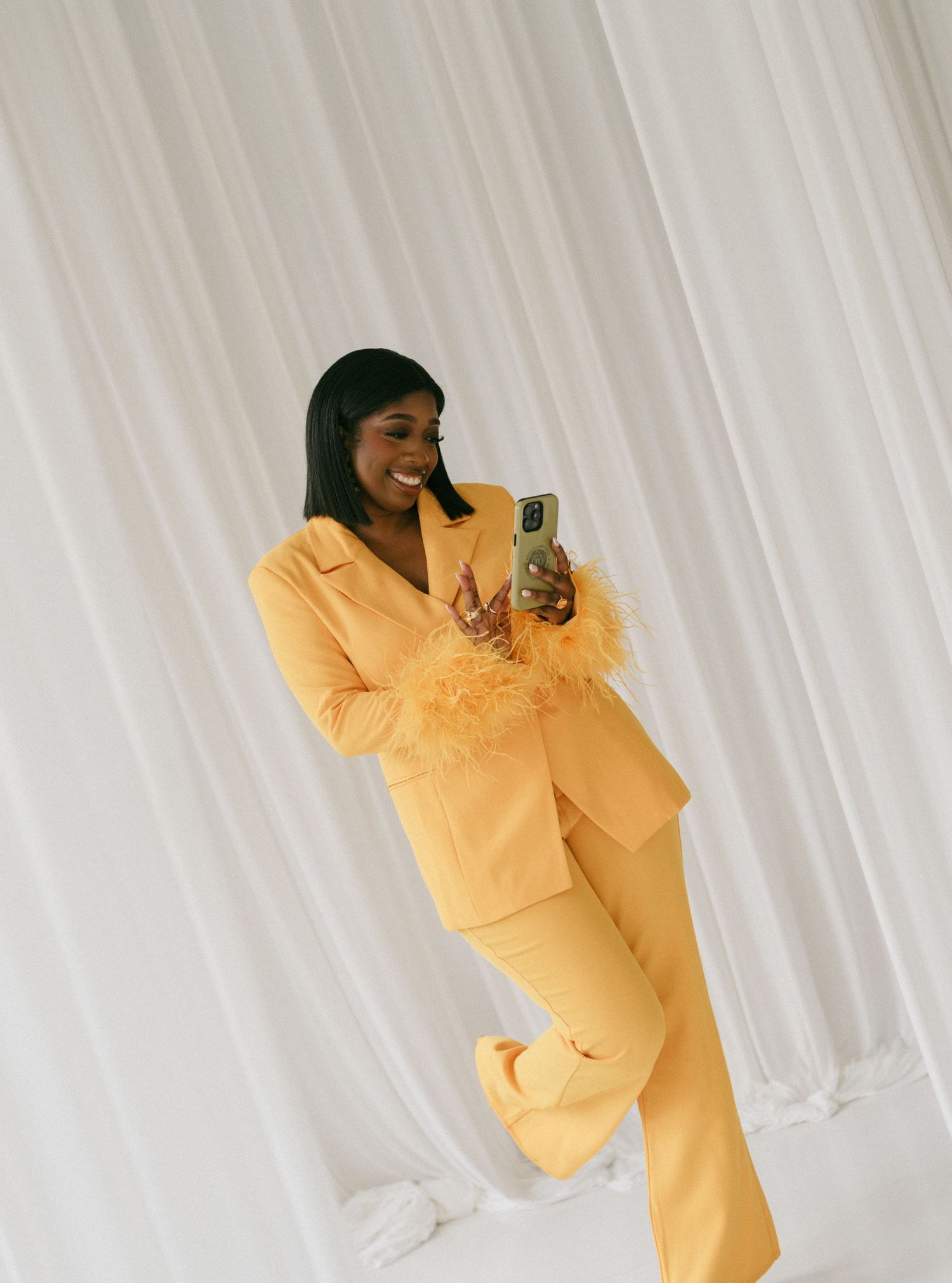 Woman in yellow feather-trimmed pantsuit taking a mirror selfie against white curtain backdrop.