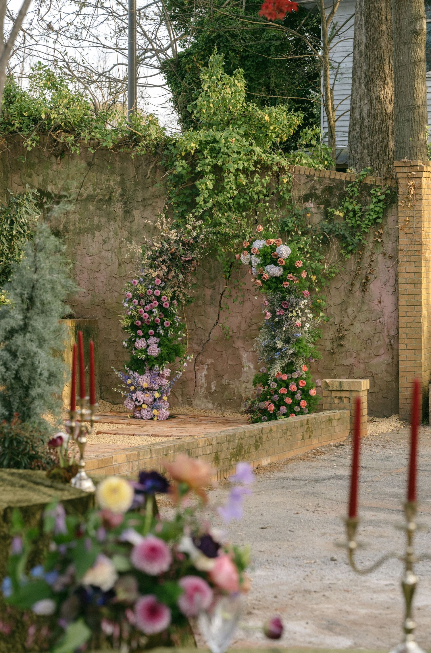 Romantic garden wedding ceremony arch adorned with pink and purple florals against a rustic stone wall.
