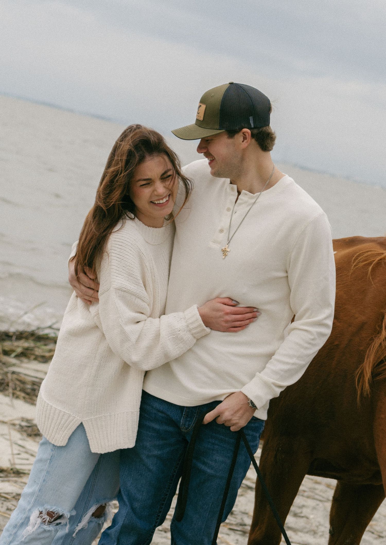 A smiling couple in cream sweaters and jeans embrace on a sandy beach near a brown horse on a cloudy day.