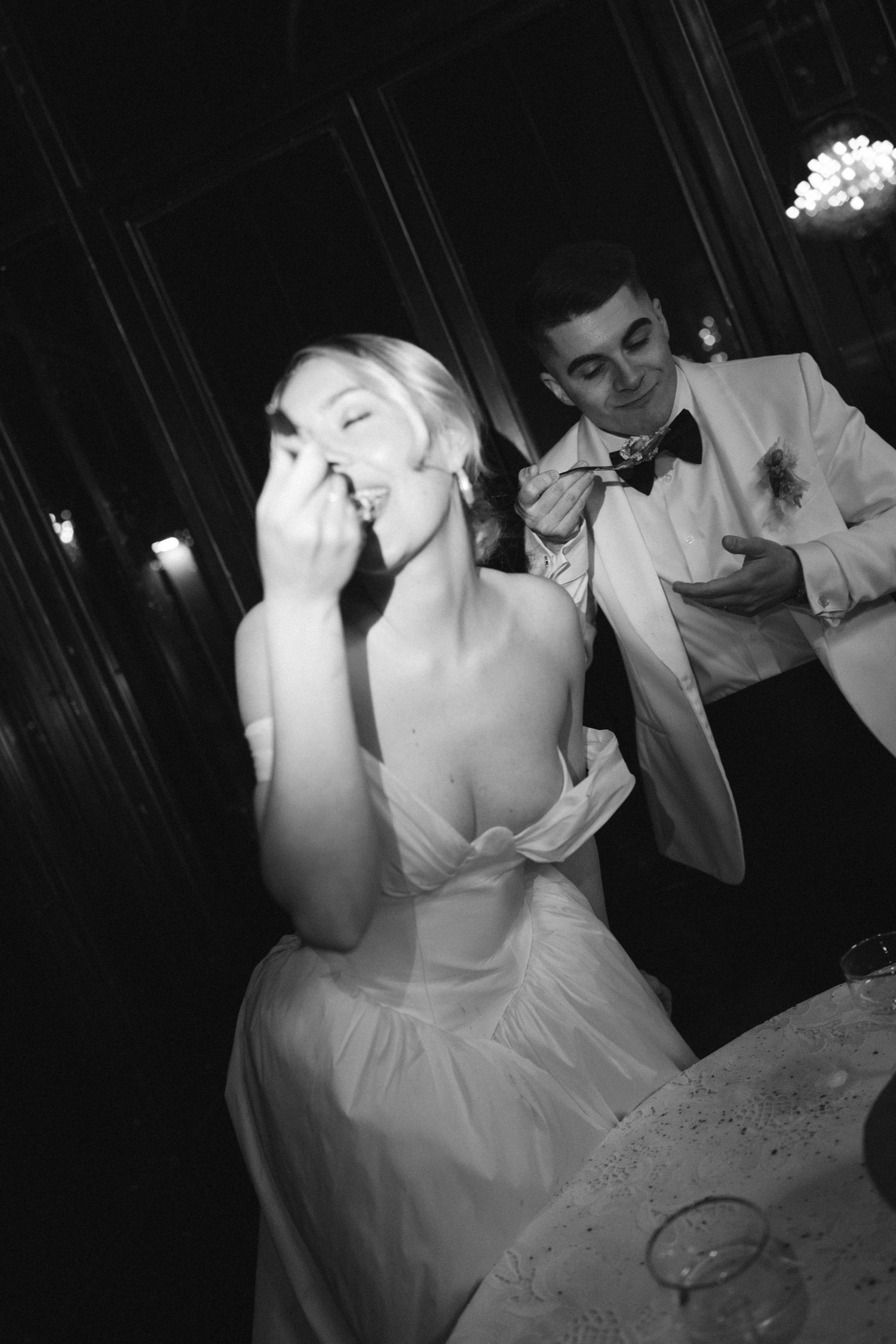 Black and white photo of a laughing bride in backless gown with smiling groom in white tuxedo at wedding reception.