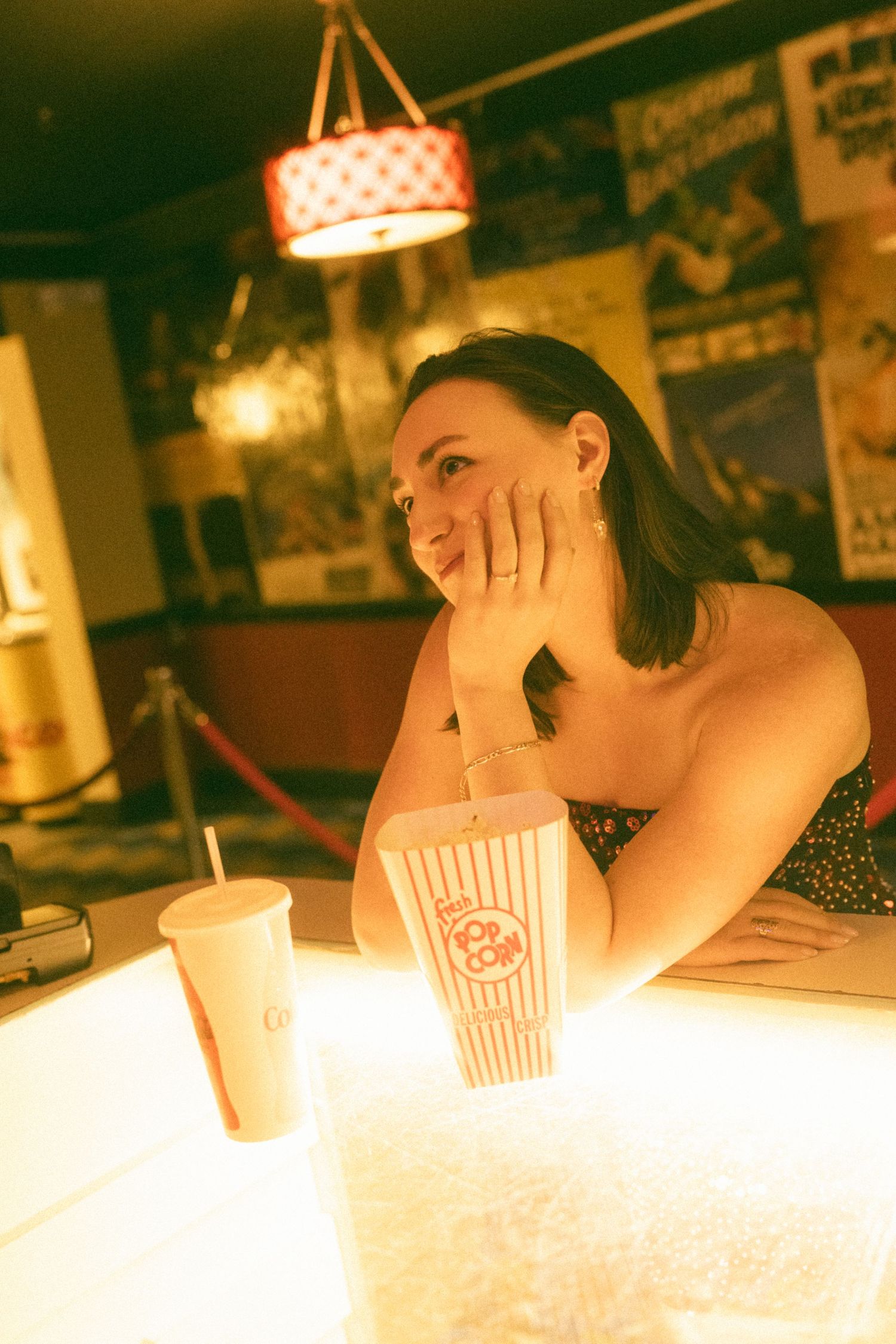 A woman rests her chin on her hand at a cinema counter with popcorn and a drink, bathed in warm golden light.