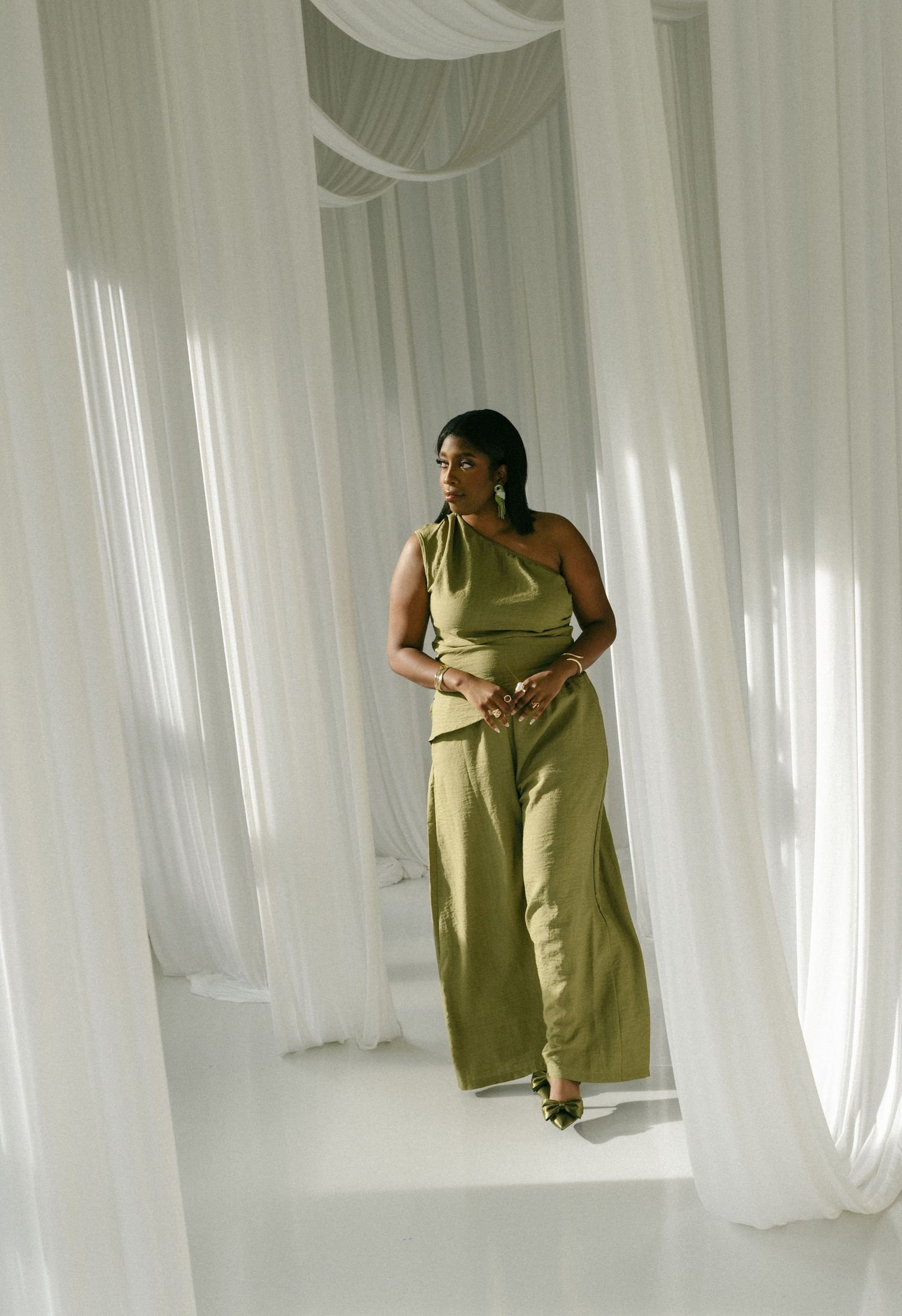 Woman in olive green satin one-shoulder jumpsuit standing among flowing white curtains in elegant setting.