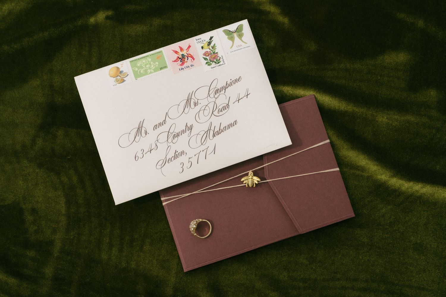 Elegant wedding invitation envelope with calligraphy address, colorful stamps, gold bee charm, and burgundy ribbon on green velvet.