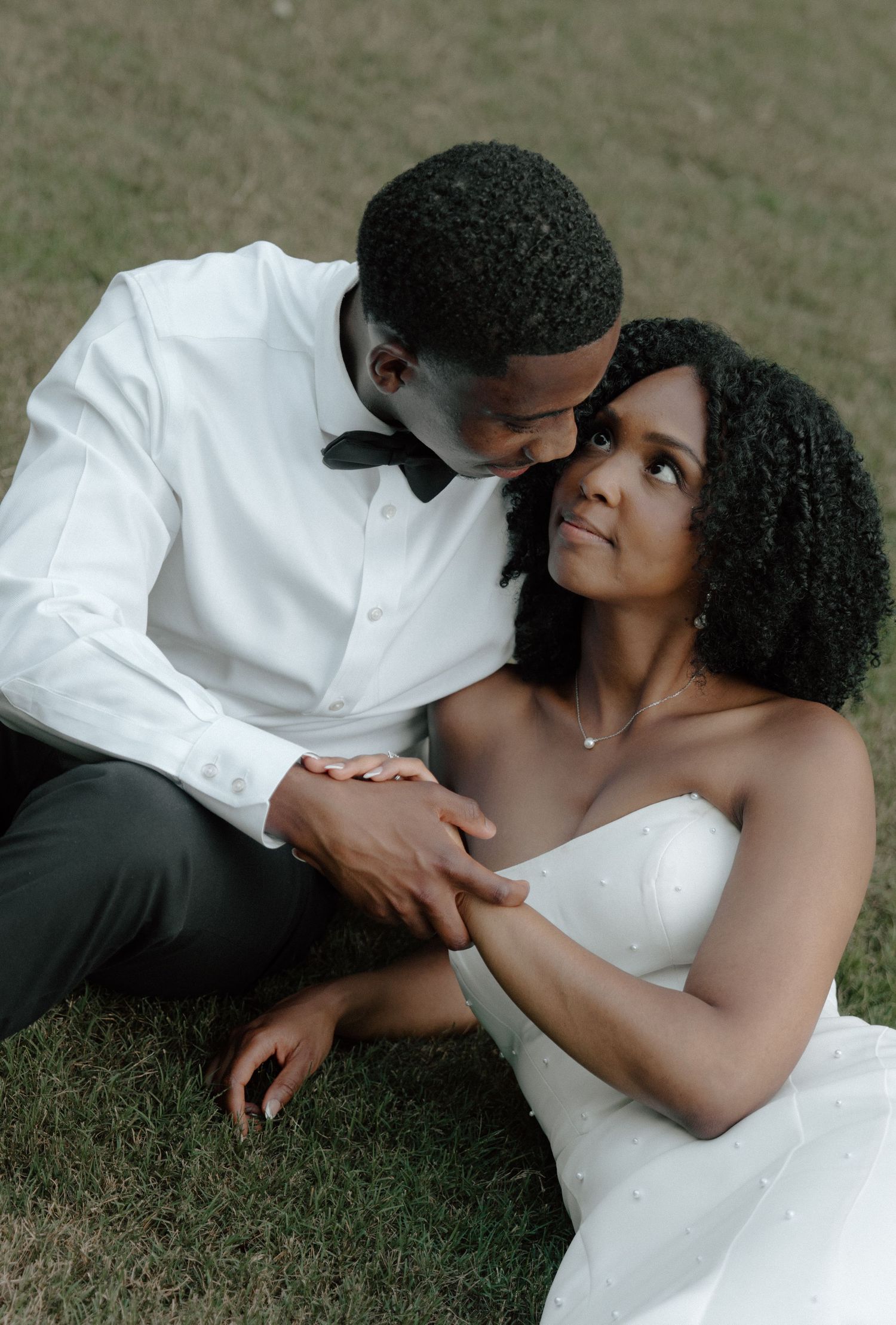 Romantic bride and groom sitting on grass, groom whispering to bride in wedding attire.