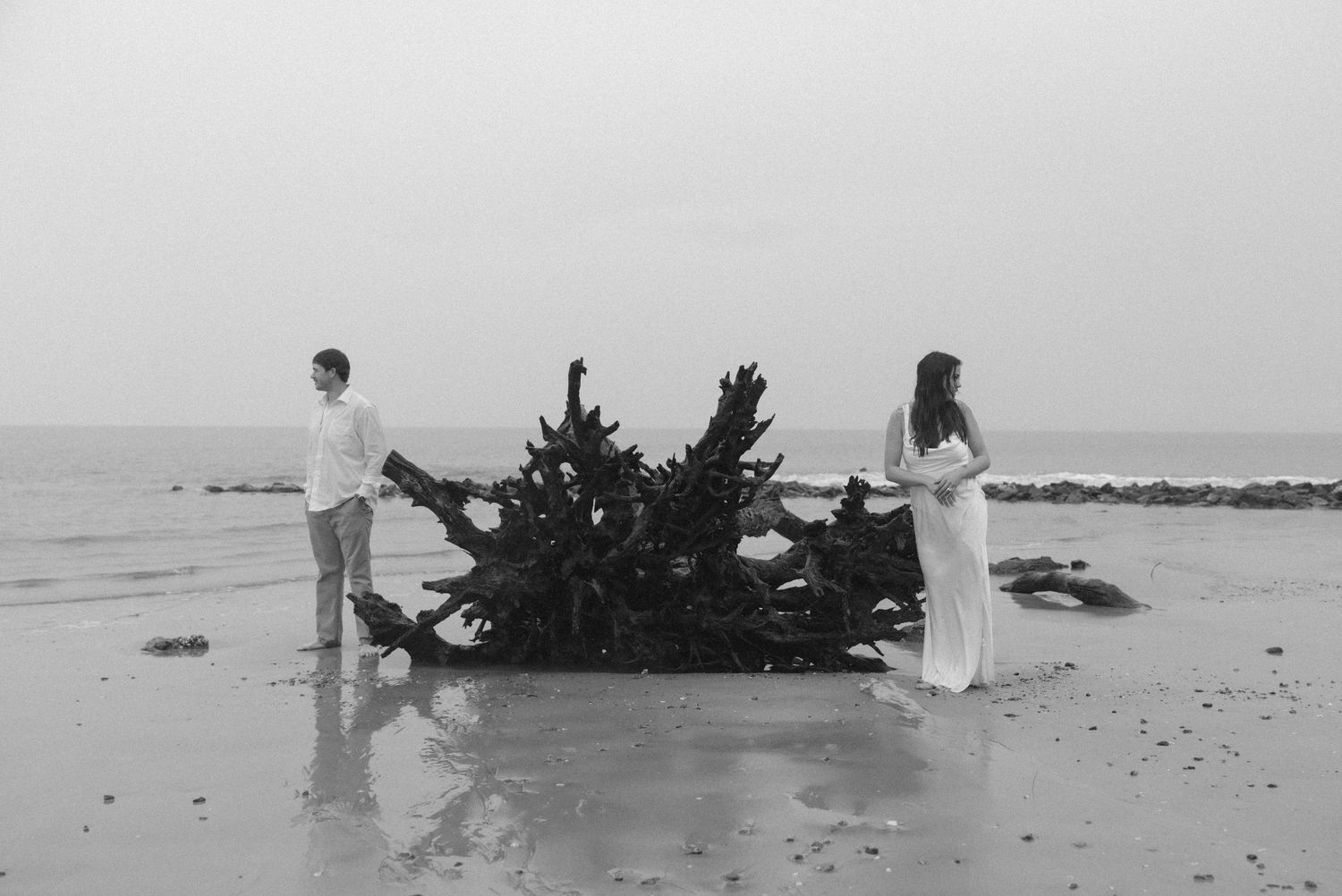 Black and white beach photo of a couple standing on either side of a large driftwood piece by the ocean.  Jekyll Island Engagement