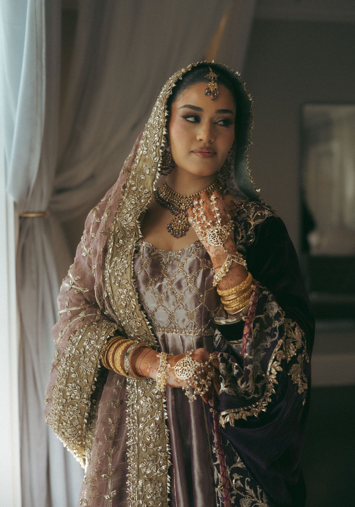 A bride in ornate gold-embroidered purple lehenga with dupatta, maang tikka, layered necklaces, and gold bangles.