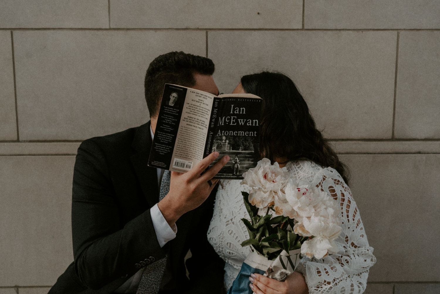 Couple hiding behind Ian McEwan book holding white peony bouquet in lace dress against stone wall.