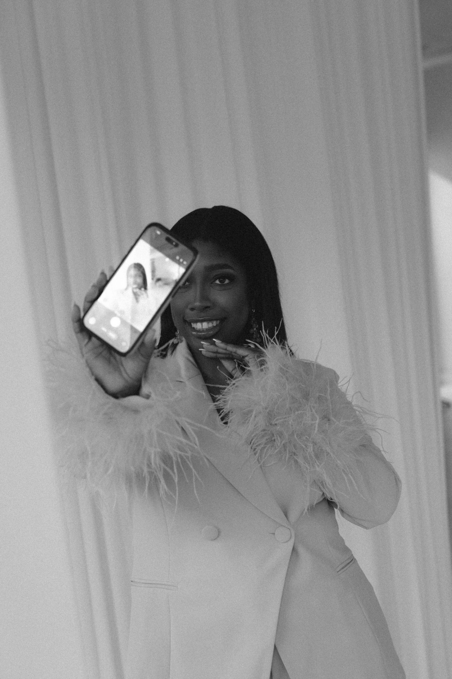 Black and white photo of a smiling woman in a feather-trimmed blazer holding up a smartphone toward the camera.