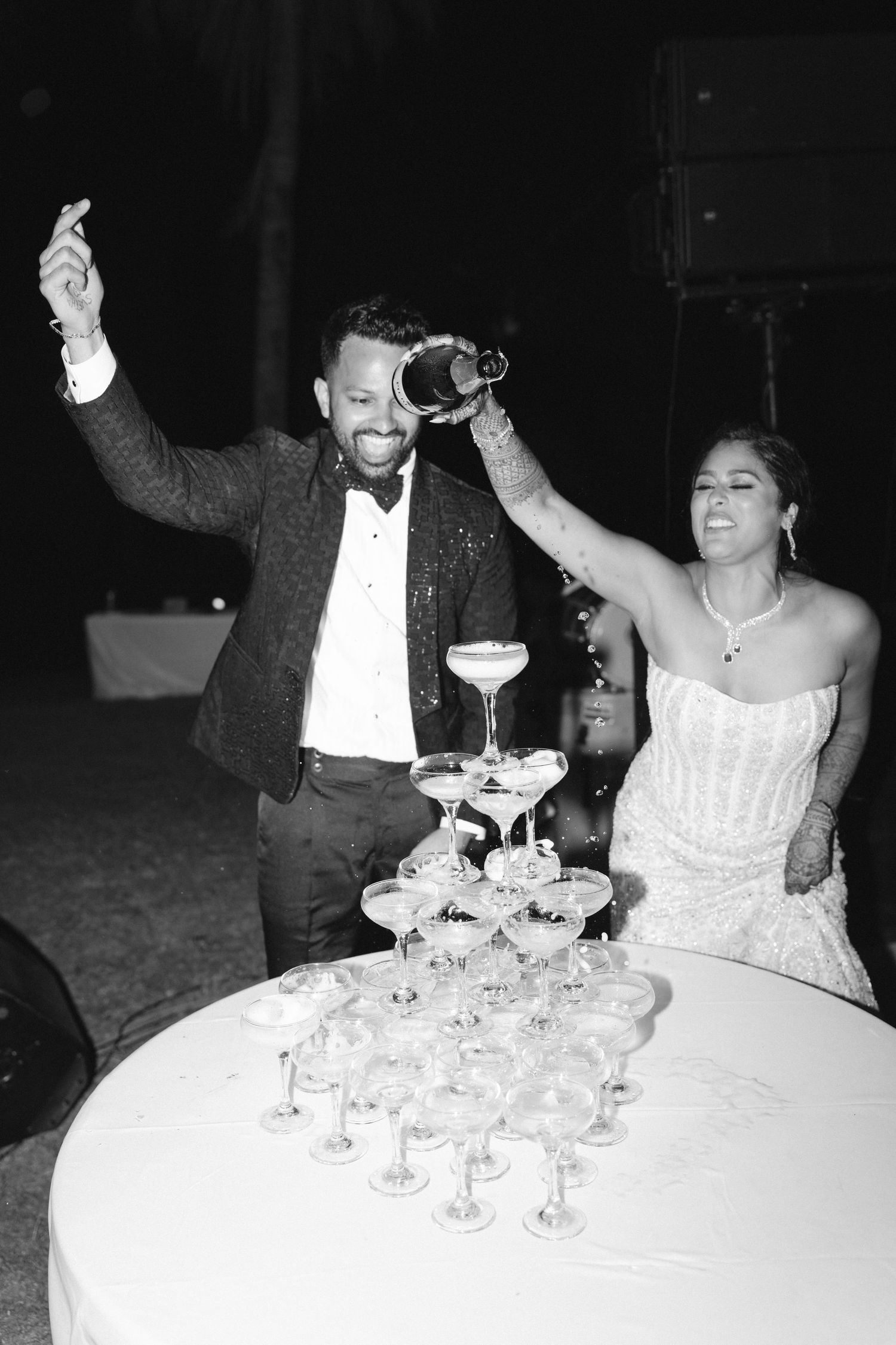 Bride and groom joyfully pouring champagne tower at wedding reception in black and white photo.