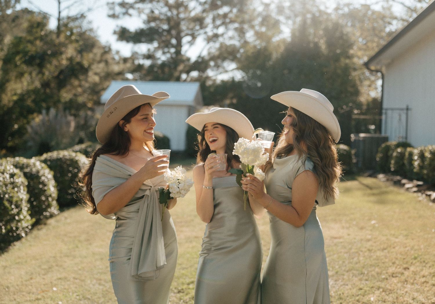 Three bridesmaids in sage green dresses and cowboy hats laughing outdoors at a wedding celebration.