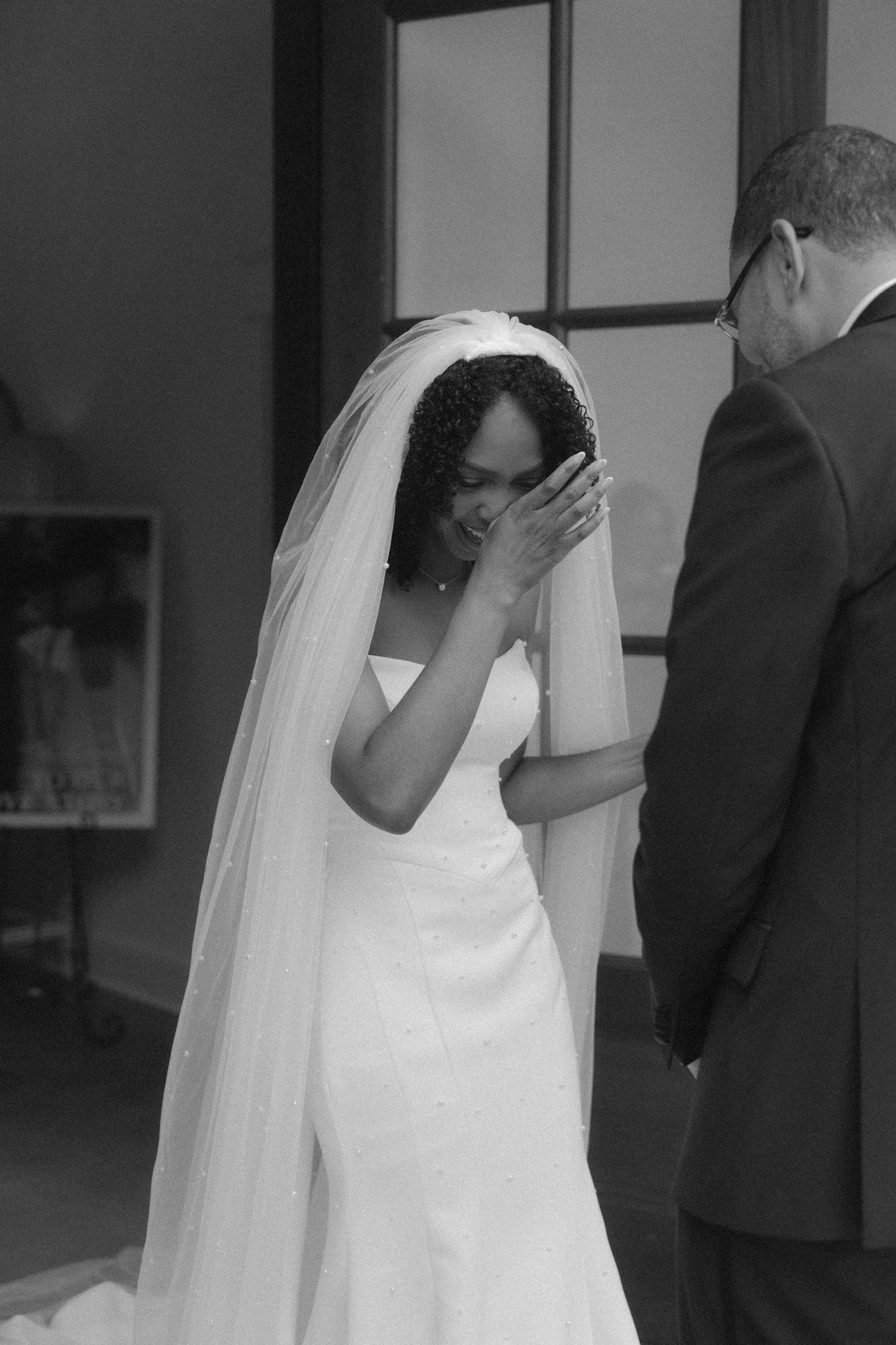 Emotional Black bride in white wedding dress and veil wiping tears during wedding ceremony.