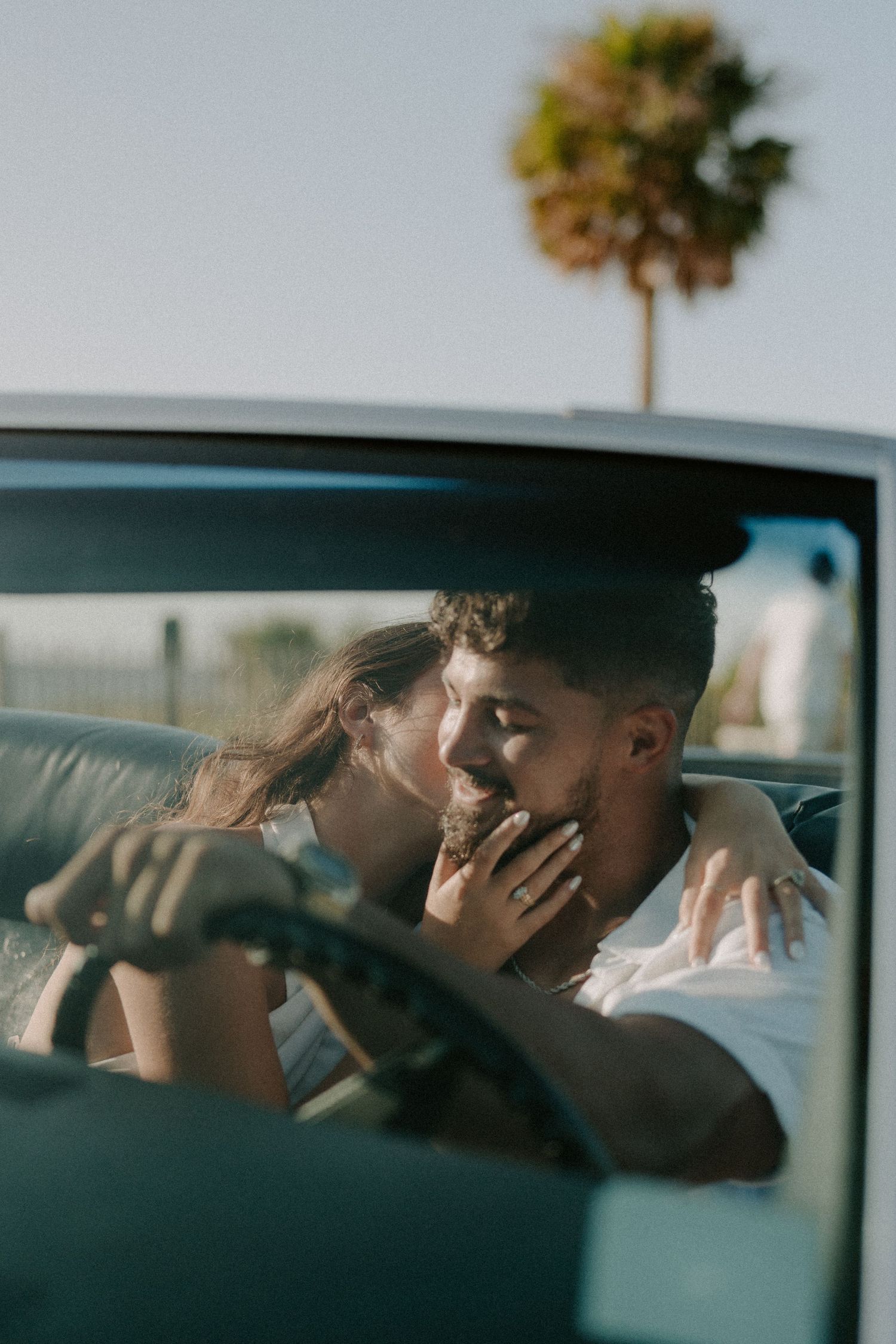 Couple sharing a romantic kiss inside a vintage car with a palm tree in the sunny background.