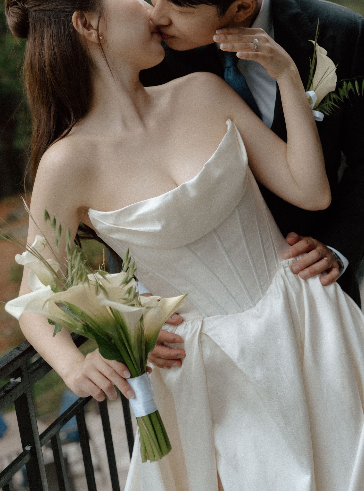 Bride in strapless white corset gown kissing groom, holding elegant calla lily bouquet with ribbon.