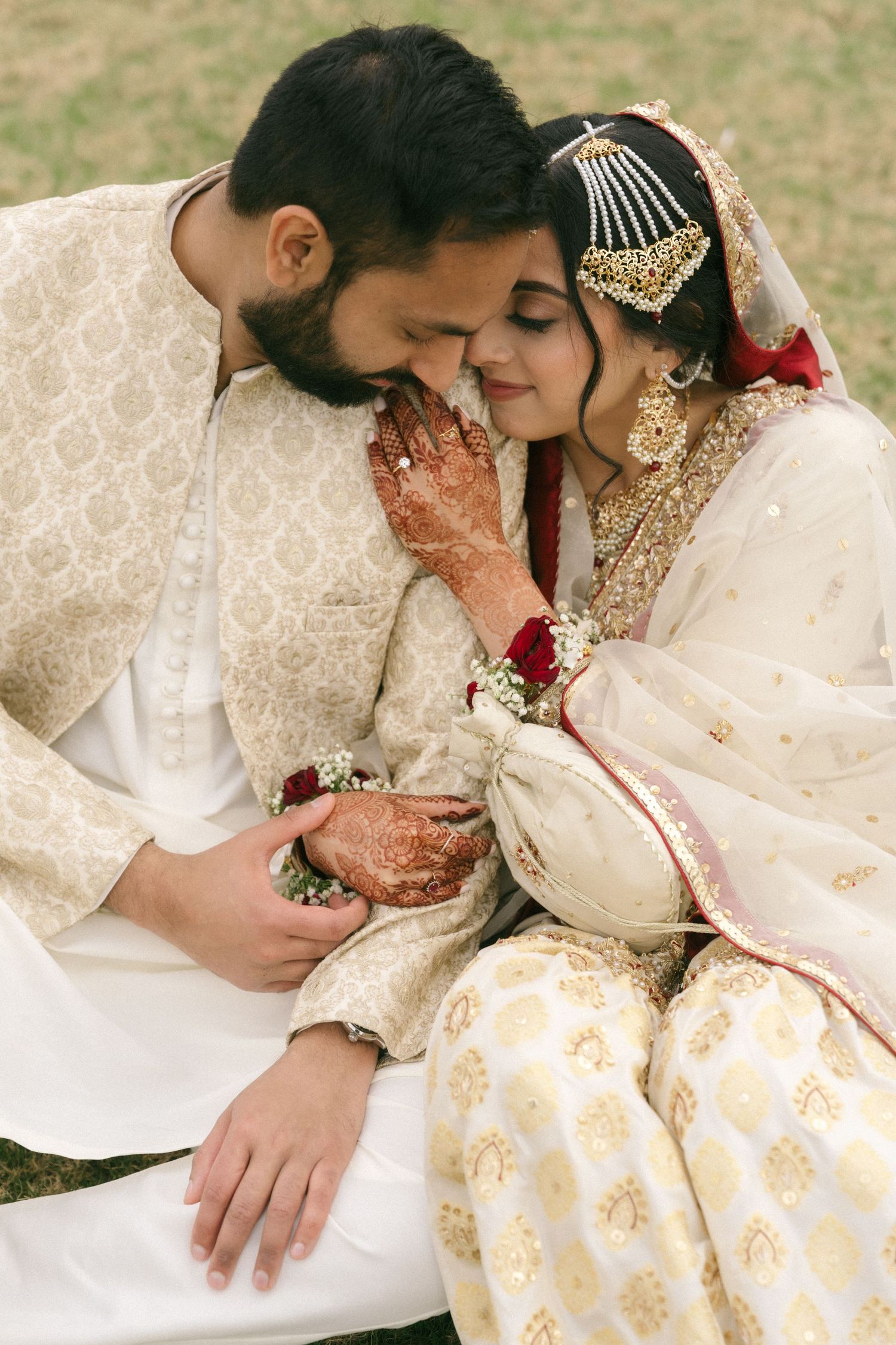 South Asian bride and groom in ivory embroidered wedding attire share a tender moment, bride adorned with mehndi and gold jewelry.