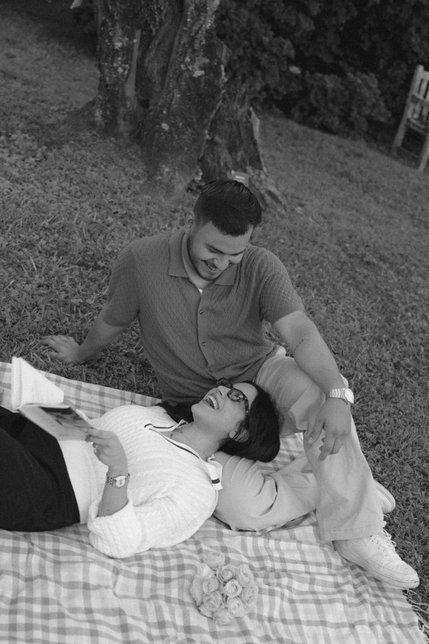 Black and white photo of a couple relaxing on a picnic blanket outdoors, woman lying down smiling up at seated man.
