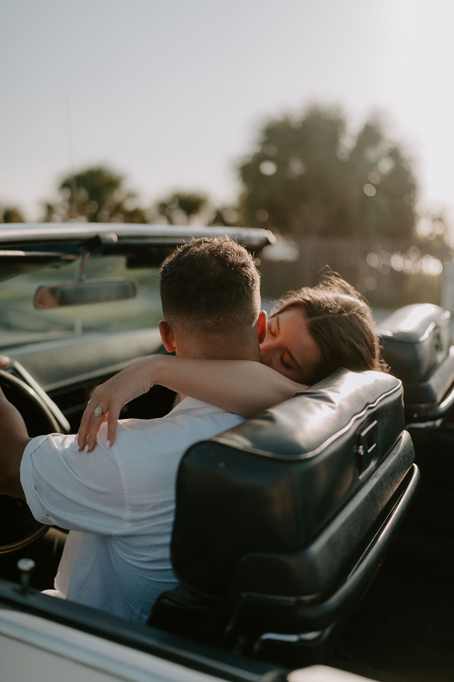 A couple embracing in a convertible car at sunset, with the woman resting her head on the man's shoulder.