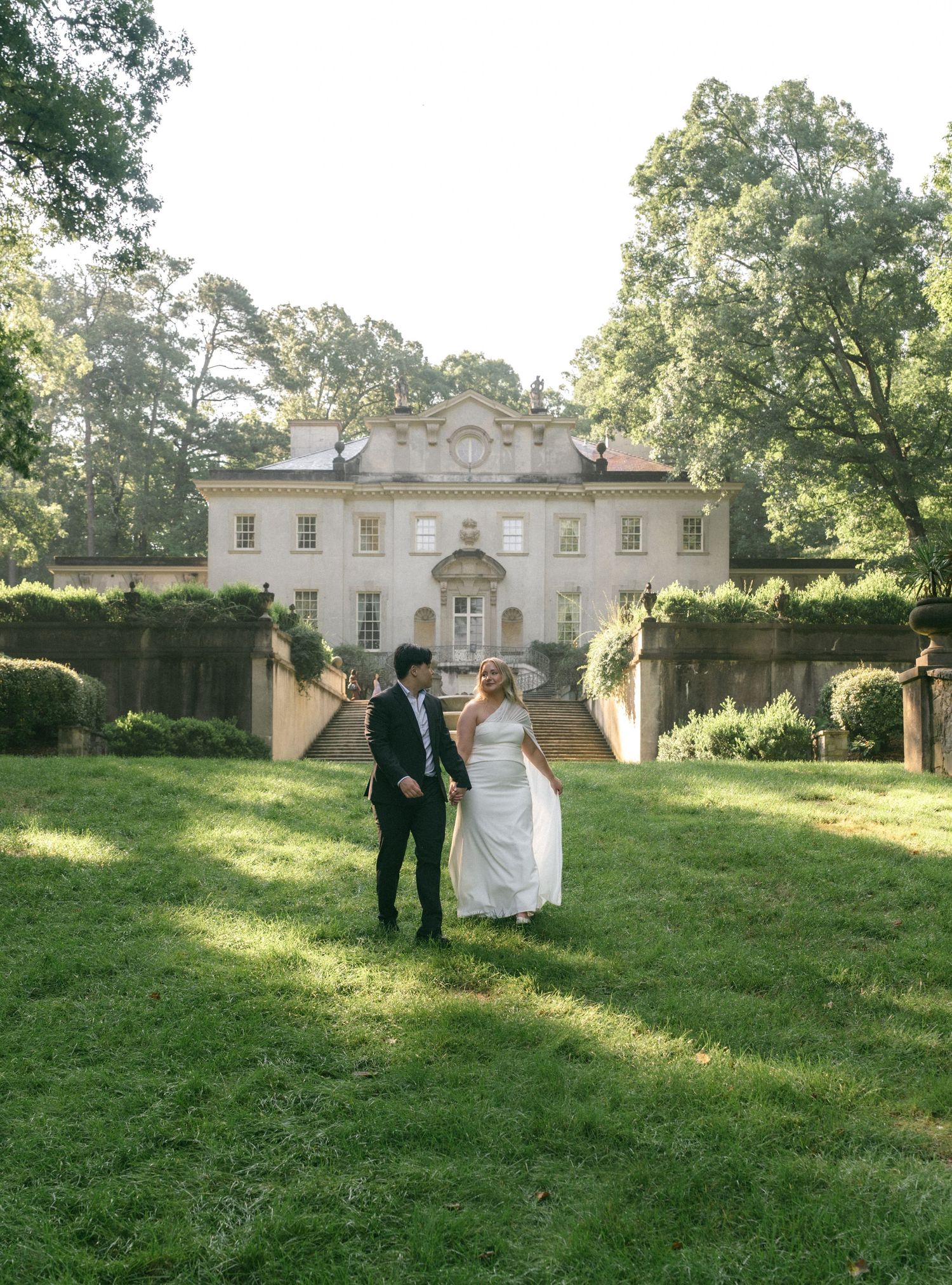 Bride and groom holding hands on lush green lawn before elegant Georgian mansion wedding venue surrounded by trees.