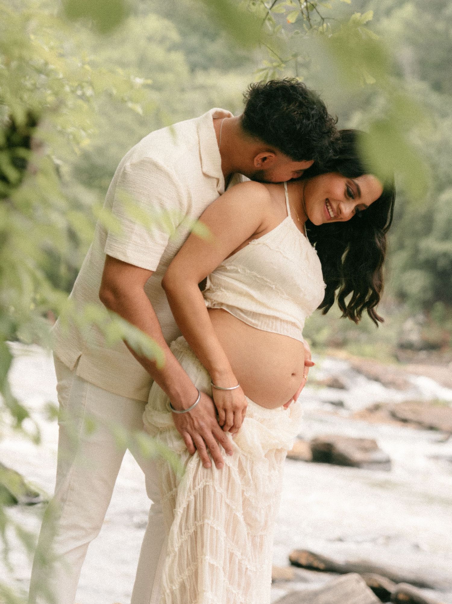 Couple in white outfits sharing a tender maternity moment outdoors by a river, woman cradling baby bump.