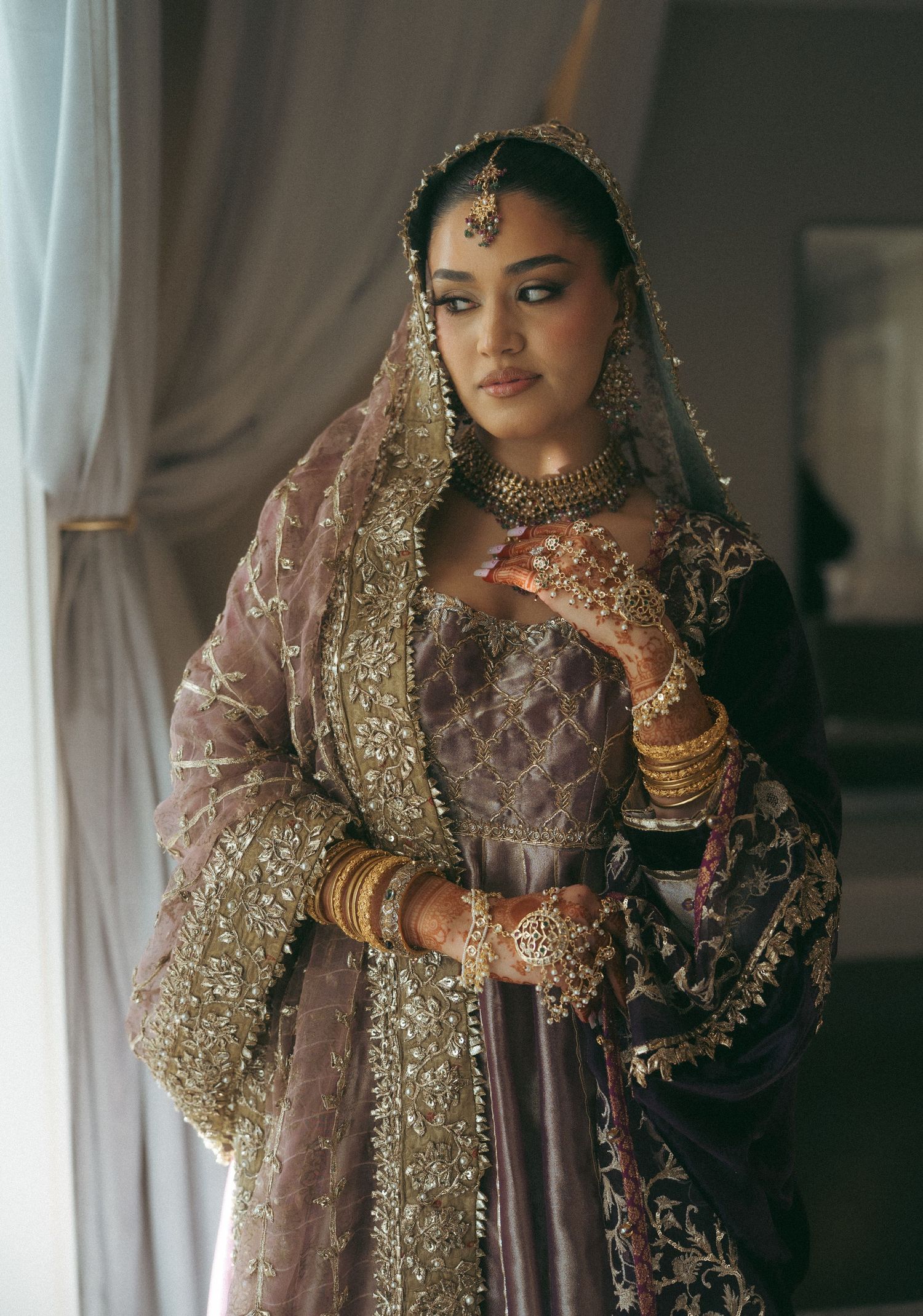A bride in ornate purple lehenga with gold embroidery, wearing traditional jewelry and dupatta, posing elegantly indoors.