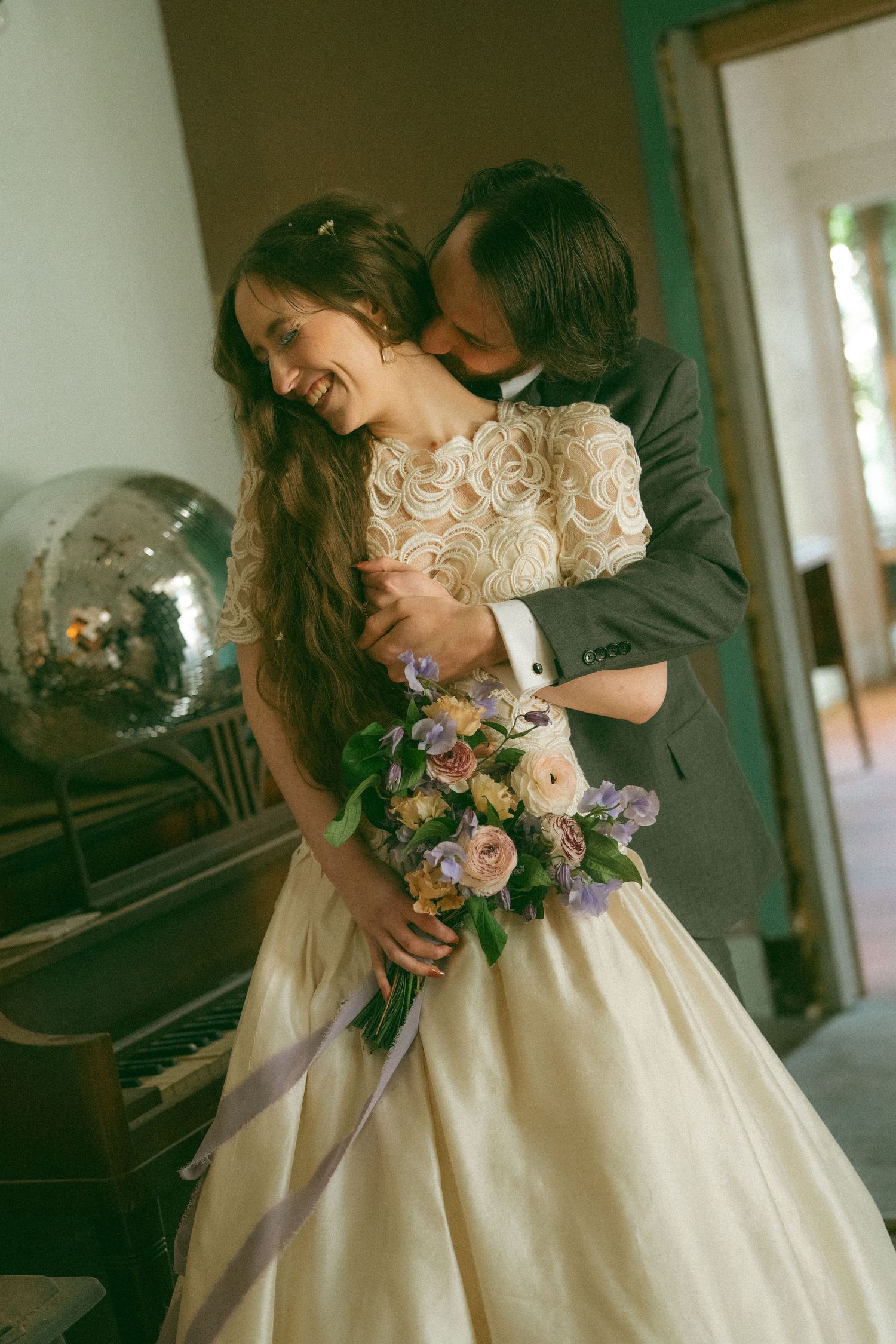 Groom embracing smiling bride holding pastel bouquet indoors near vintage piano and disco ball.