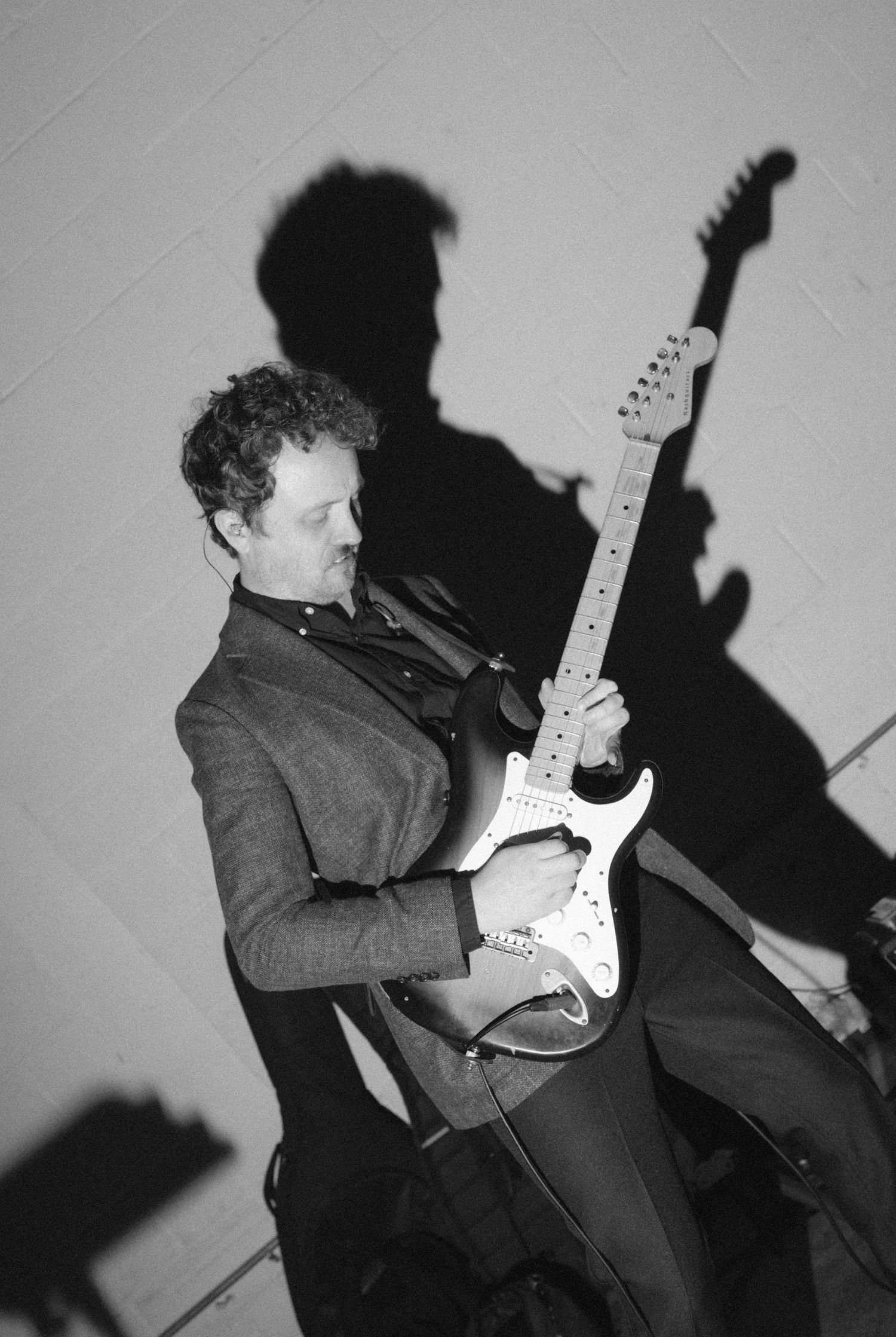 Black and white photo of a man playing electric guitar, casting a dramatic shadow on the wall behind him.