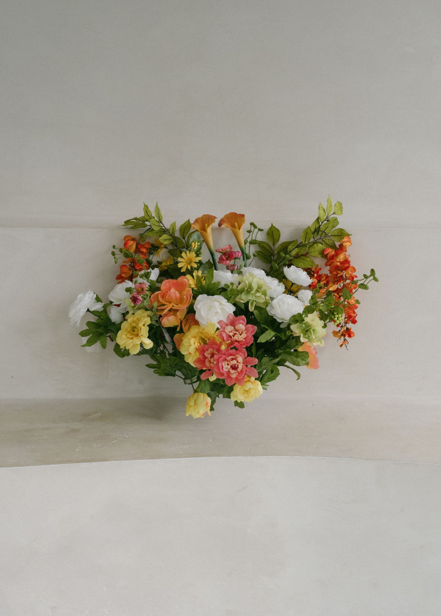 Colorful floral arrangement with orange, pink, white, and yellow blooms and lush green foliage on a neutral background.