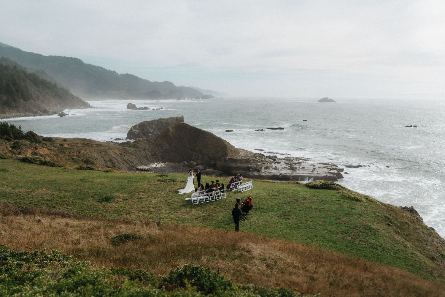 Aerial view of a wedding ceremony on a coastal cliff overlooking the Pacific Ocean on a misty day.