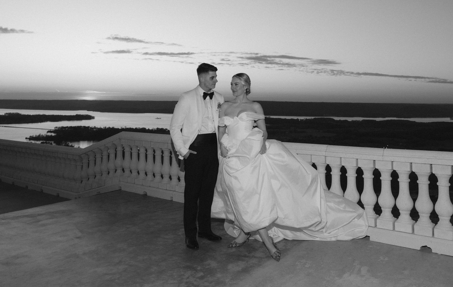 Black and white wedding photo of bride and groom on balcony at sunset overlooking water.