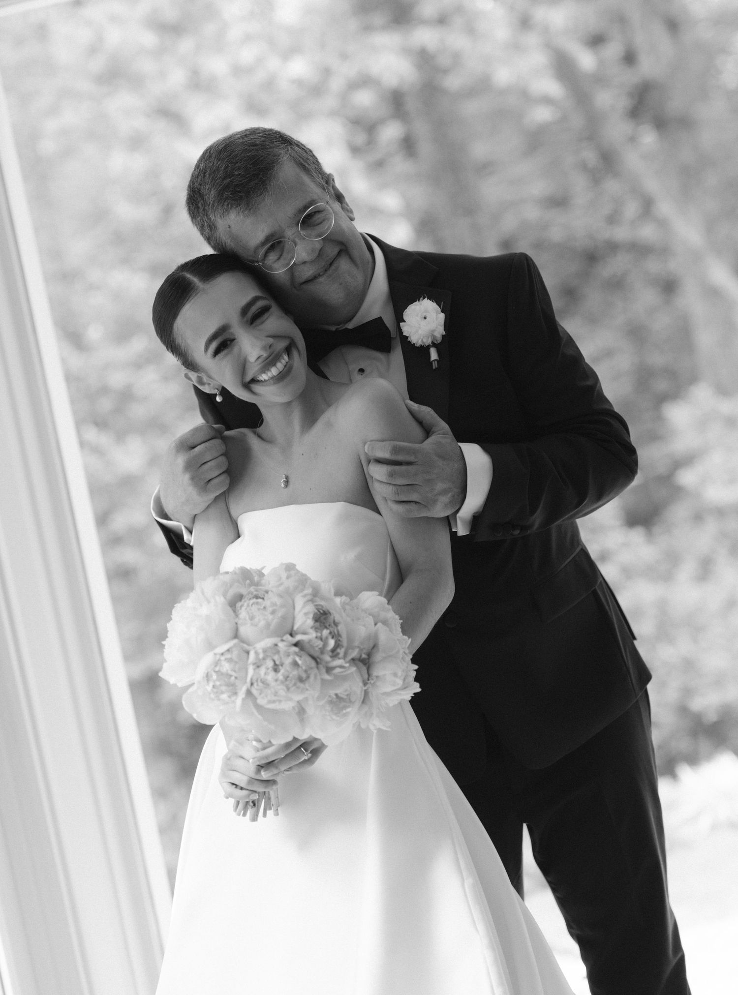 Black and white wedding photo of smiling bride holding bouquet embraced by groom in tuxedo with boutonniere.
