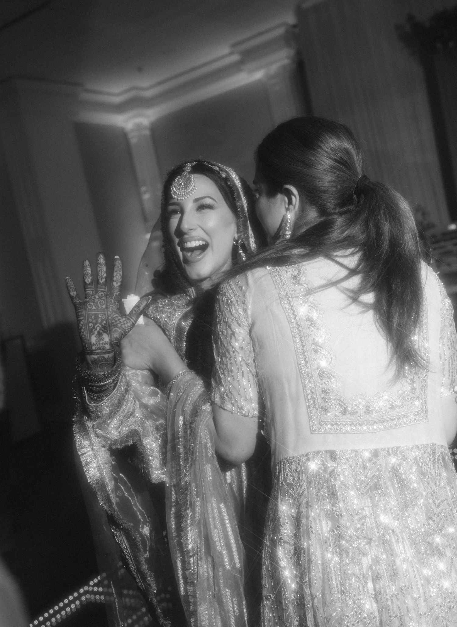 Black and white photo of a laughing South Asian bride in traditional attire with mehndi, sharing a joyful moment with a guest.