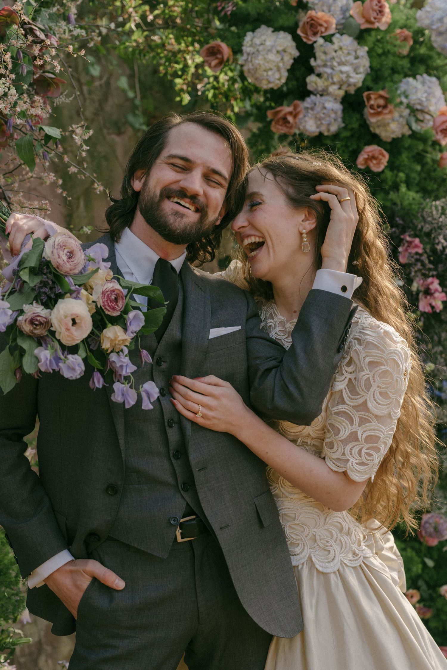 Smiling bride and groom laugh together in front of a floral arch with pink and white blooms.