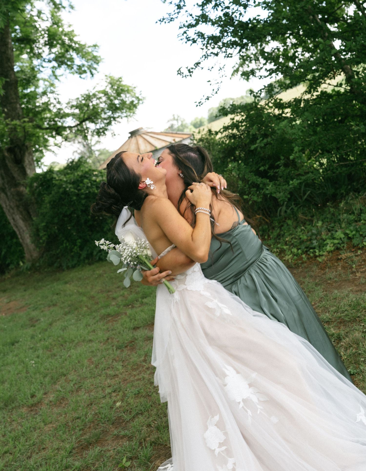 Bride in white gown embracing bridesmaid in sage green dress outdoors, sharing a joyful hug at a wedding celebration.