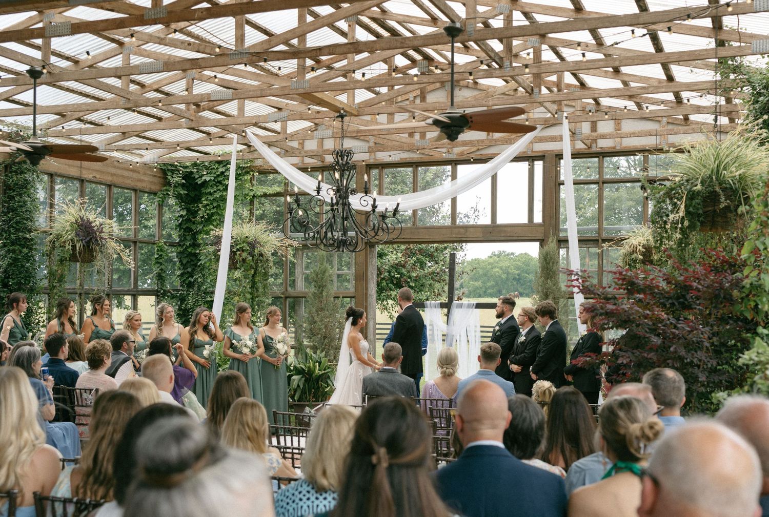 Wedding ceremony in a greenhouse venue with ivy walls, chandelier, and guests seated as couple stands at the altar.
