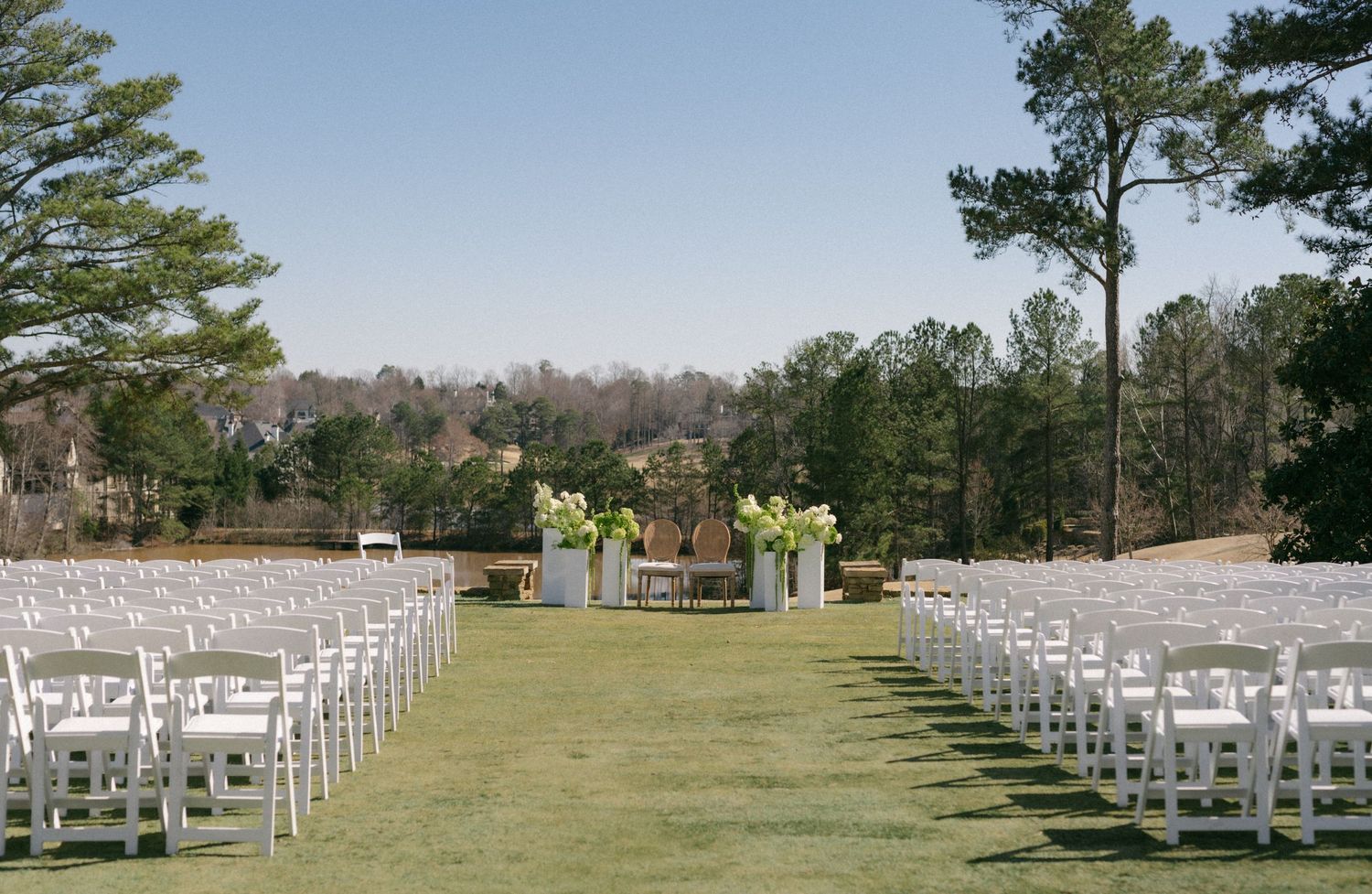 Outdoor wedding ceremony setup with white folding chairs on green grass, floral arrangements, and tall trees under a clear blue sky.
