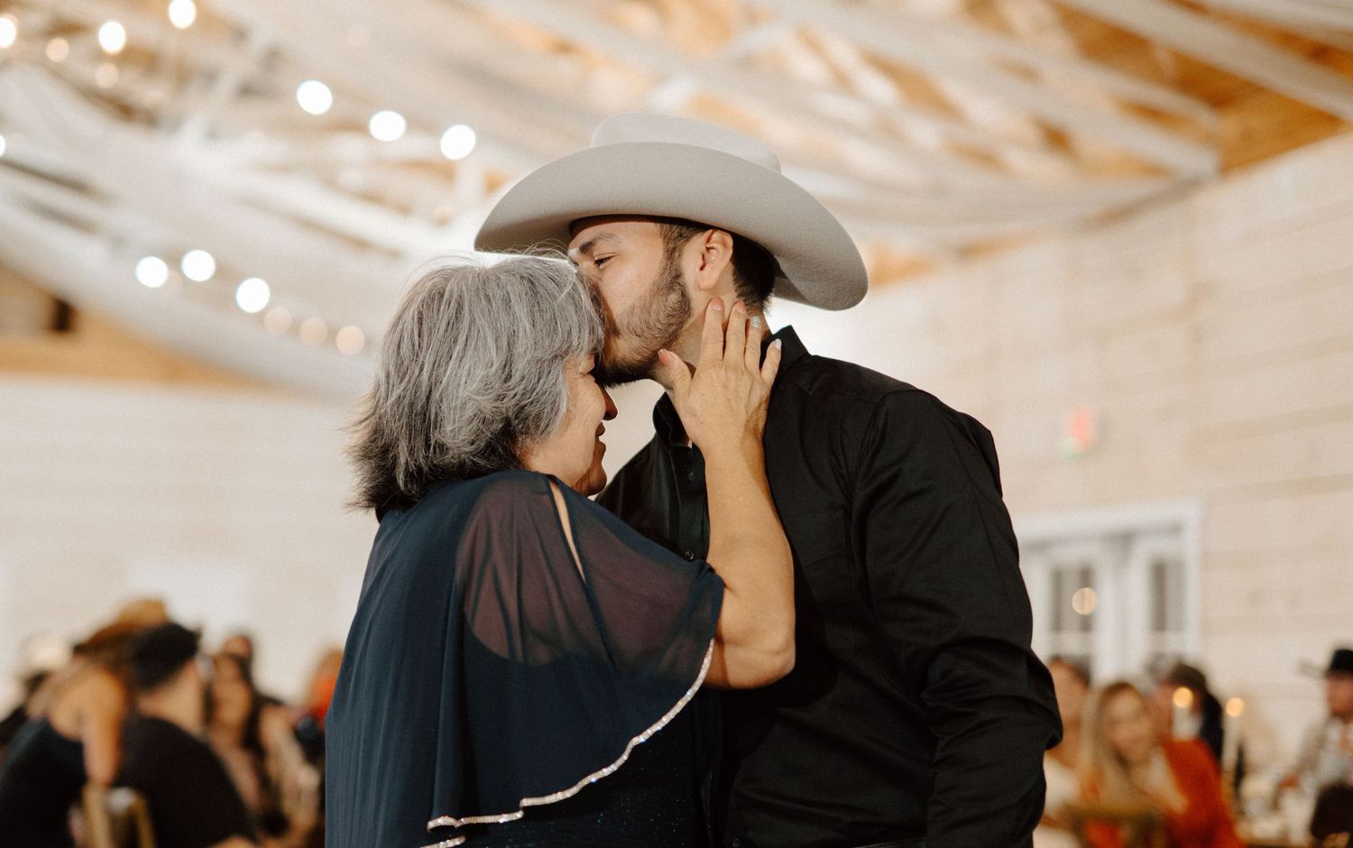 A man in a black shirt and cowboy hat shares a tender kiss with an older woman at a rustic wedding reception.