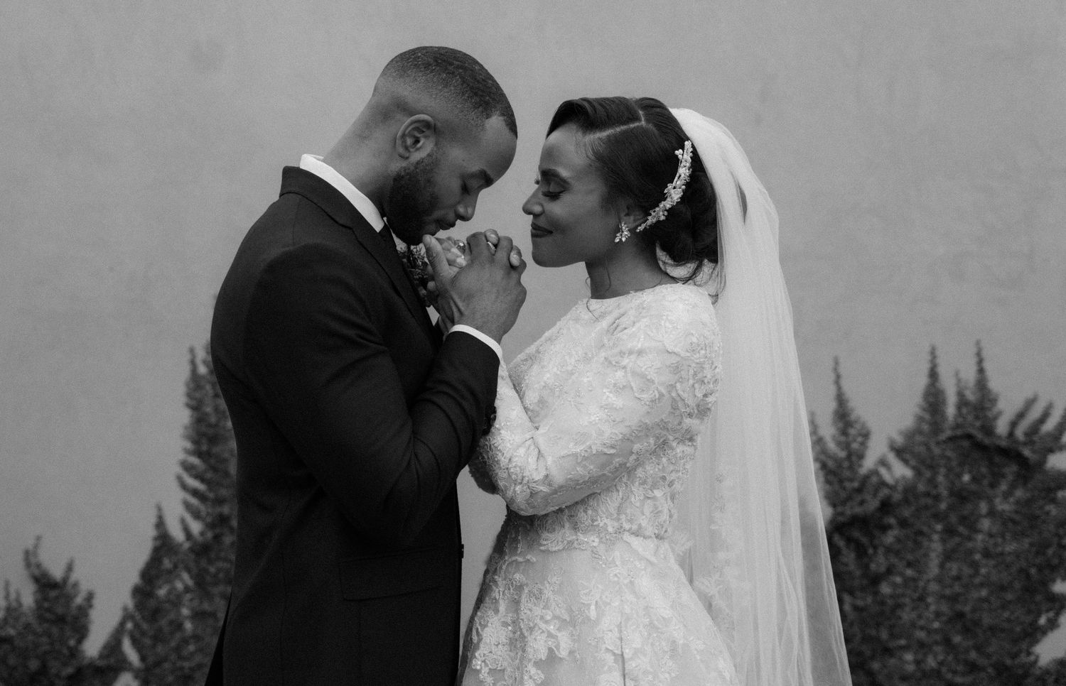 Black and white wedding photo of a couple in formal attire, groom kissing bride's hands outdoors.