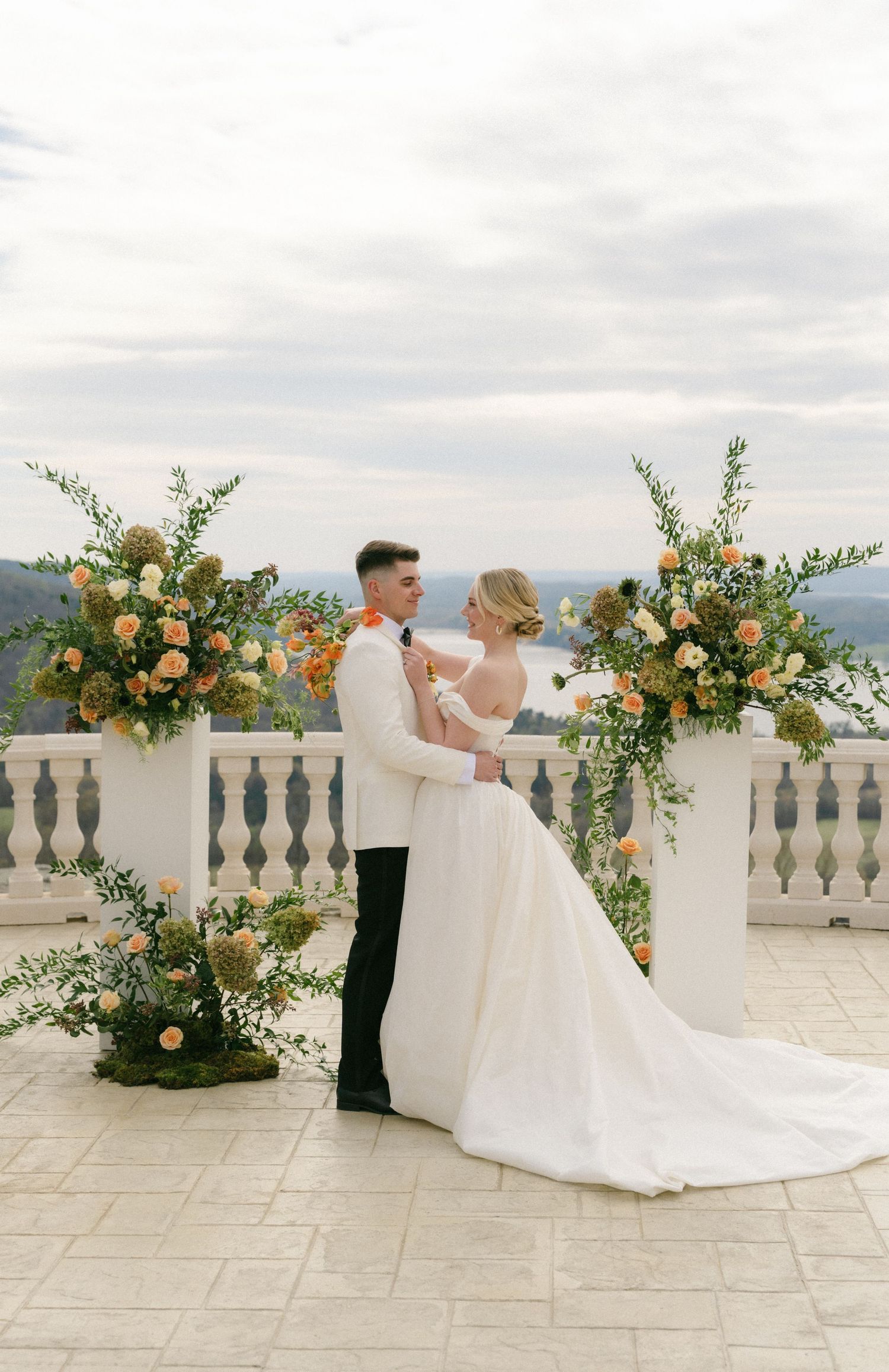 Bride and groom share a romantic moment at an outdoor wedding ceremony with lush floral arrangements on a stone terrace.