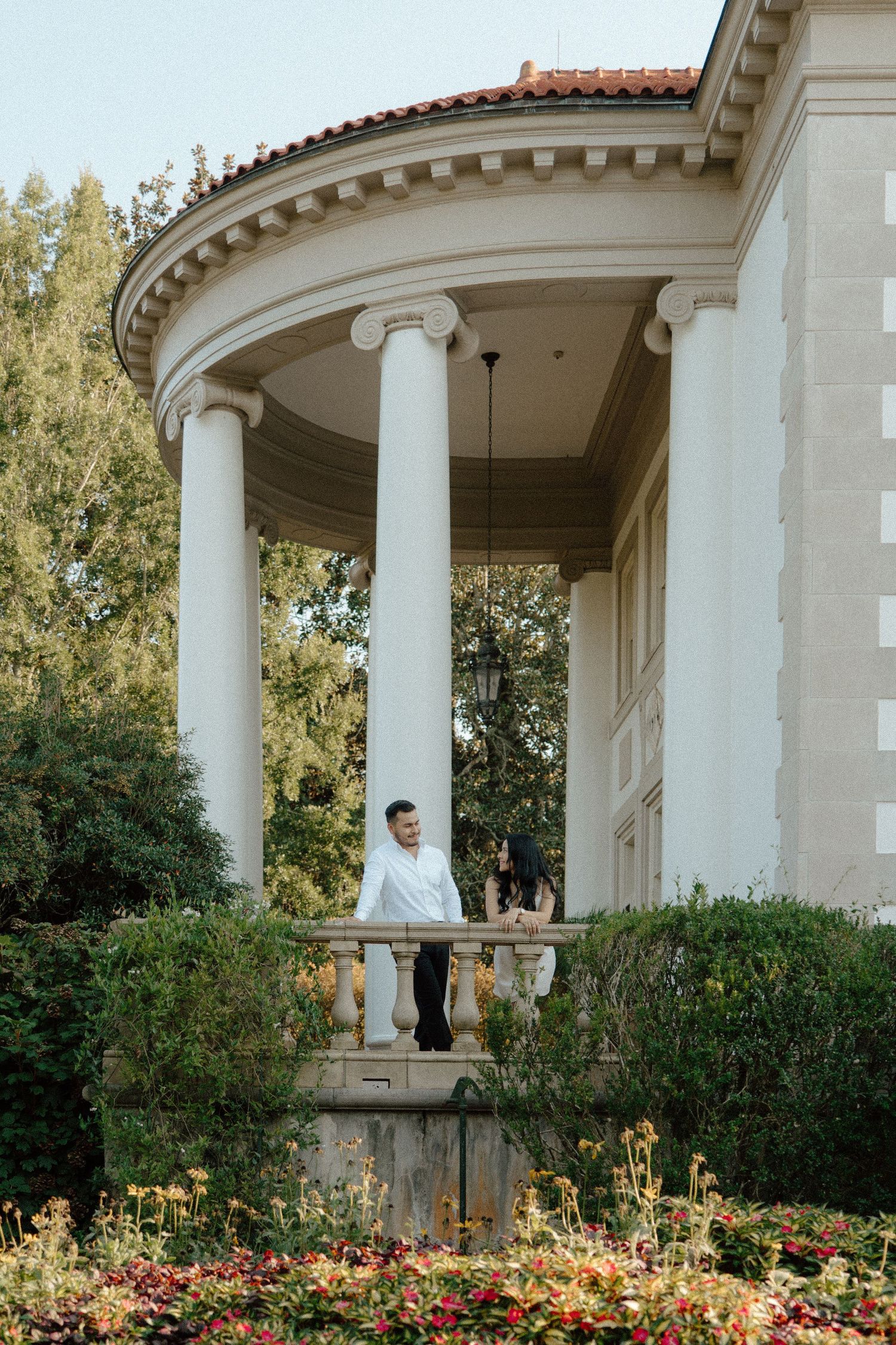 Couple standing on elegant columned portico of classical white mansion surrounded by lush gardens and colorful flowers.