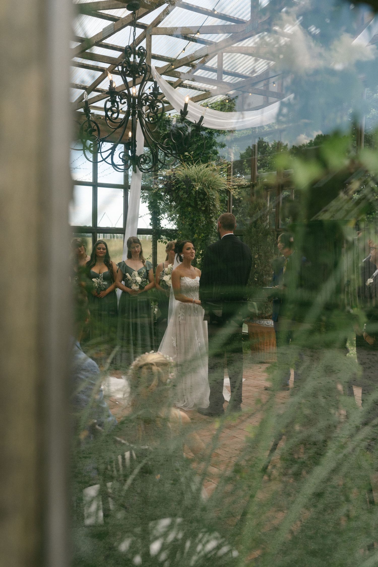 A bride and groom exchange vows in a lush greenhouse wedding ceremony with bridesmaids standing nearby.