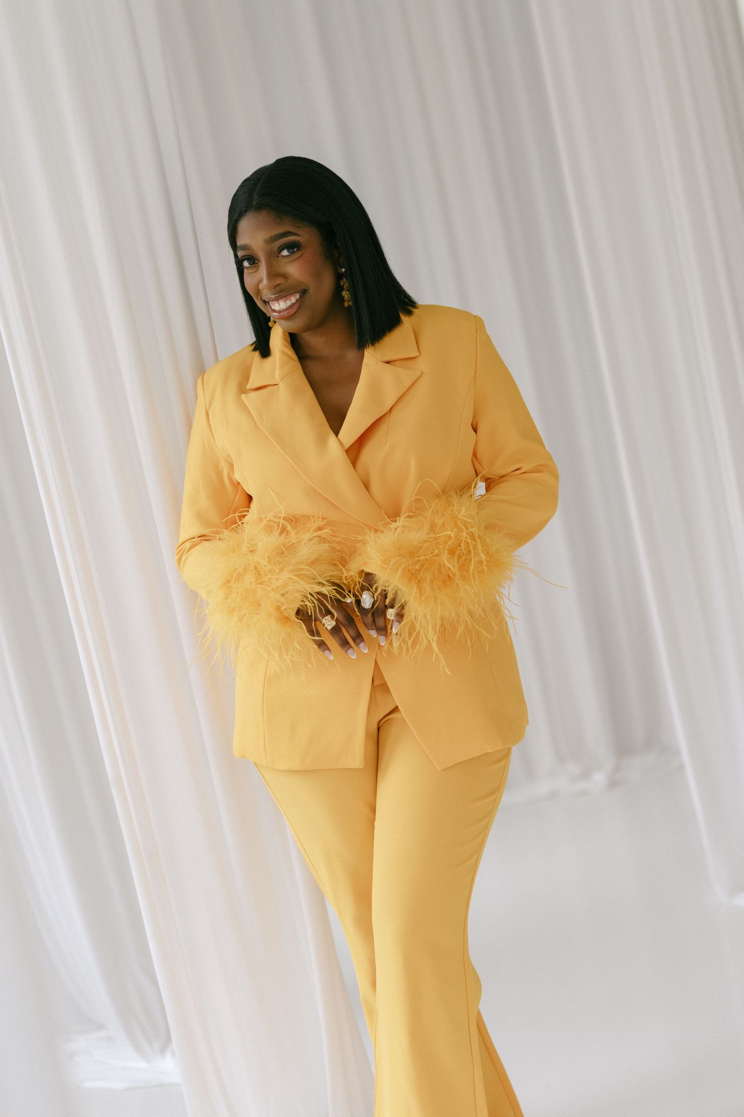 Woman in yellow feather-cuffed blazer and flare pants suit, smiling against white curtain backdrop.