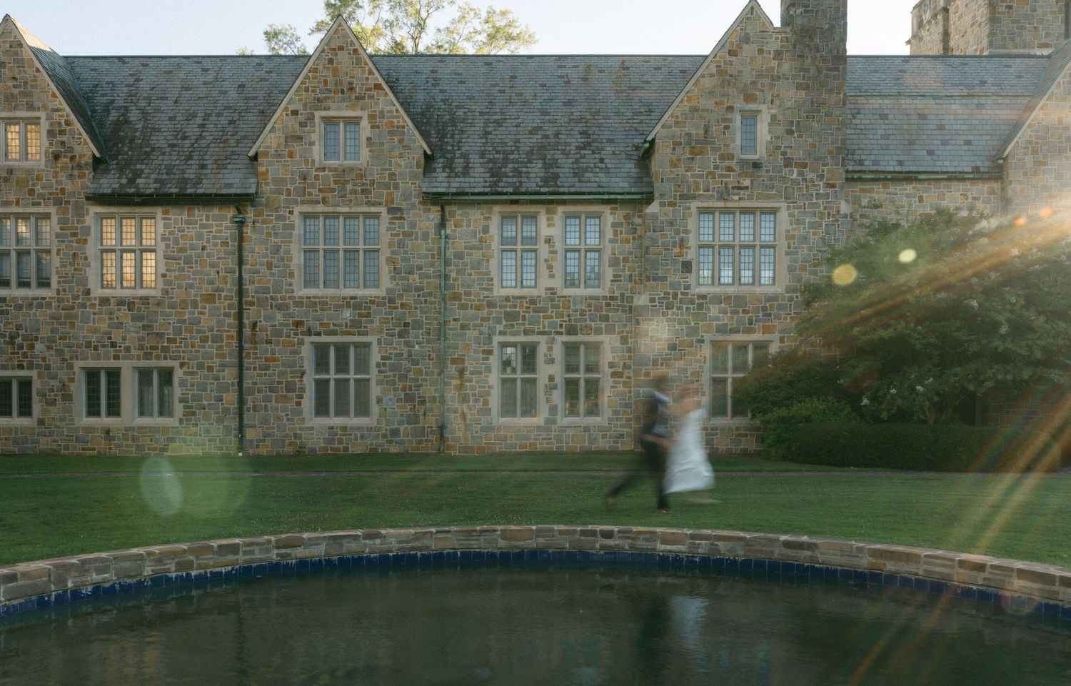 A couple dances near a reflecting pool outside a grand stone manor house surrounded by lush green lawns at dusk.